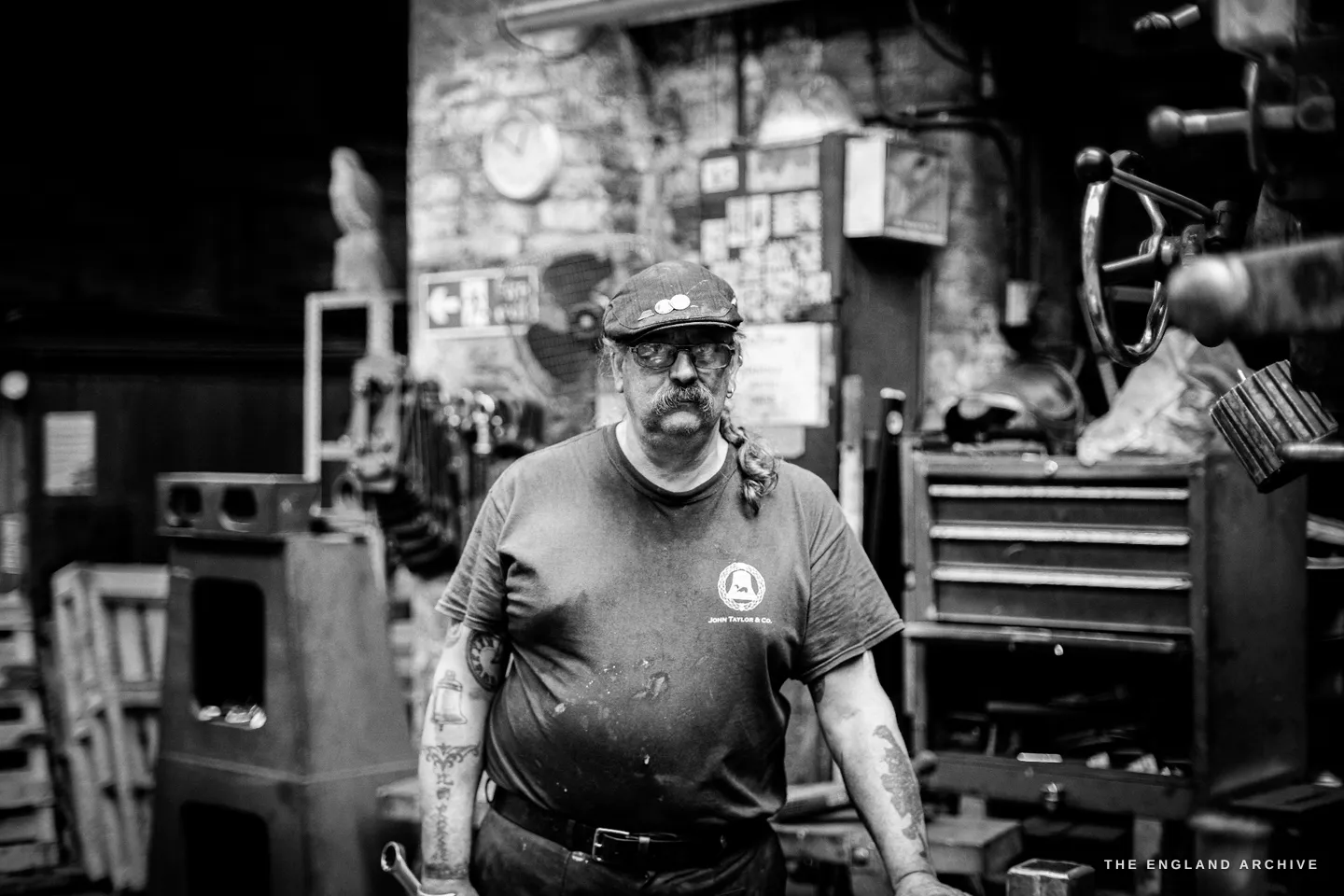 Bill standing in his brick-walled workshop, a steering-wheel-shaped machine handle to his right, a clock and pinned papers on the wall, a tool chest beside him.