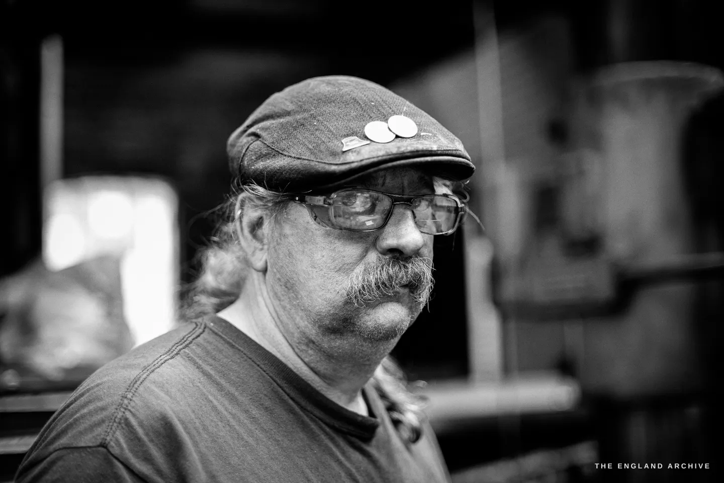 Close portrait of Bill Bowes in three-quarter profile, a flat cap with two enamel badges pinned to it, wire-framed glasses, a heavy grey moustache, the foundry dark behind him.