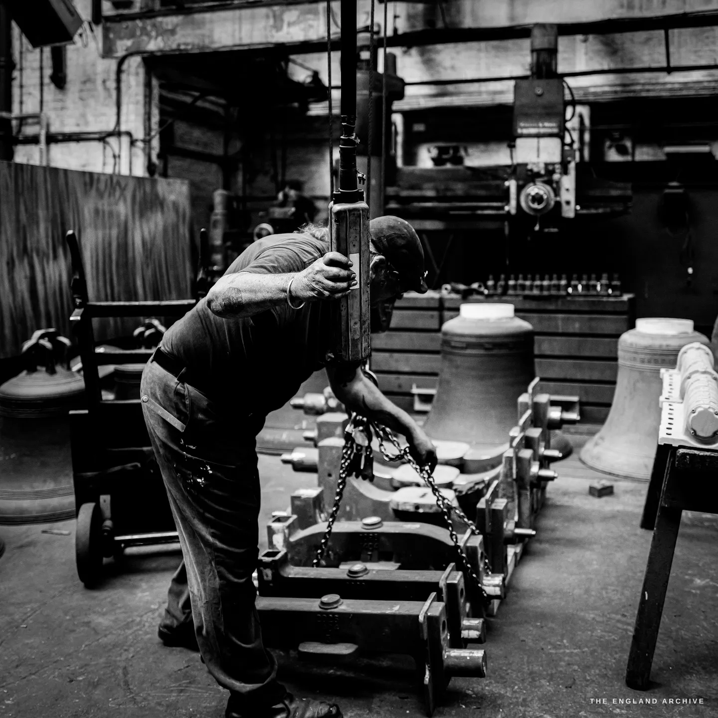 Bill bent over a long steel headstock frame, both hands hauling on a chain hoist that runs up to a beam, bells ranged on the floor around him.