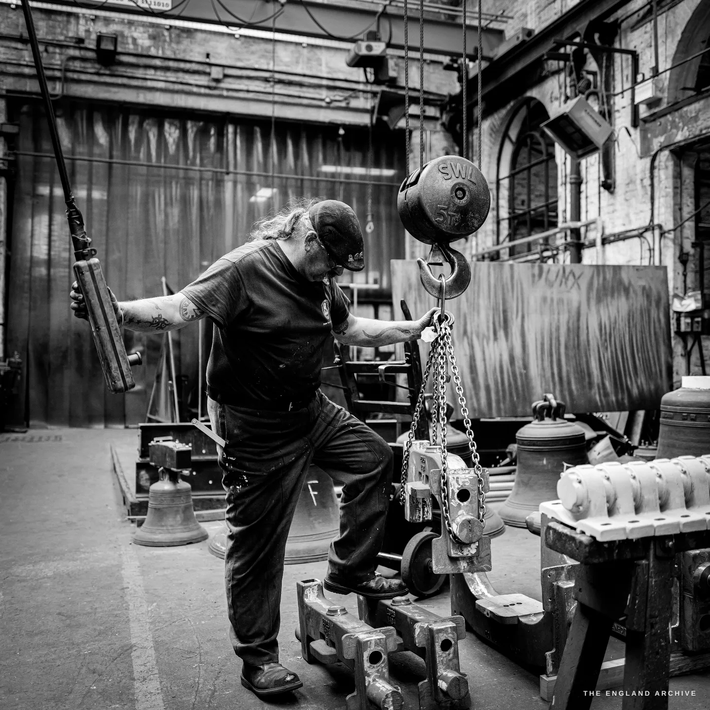 Bill with one boot up on a pallet truck, steadying a chain hoist whose load gauge reads its safe working limit, a cast frame slung beneath it.