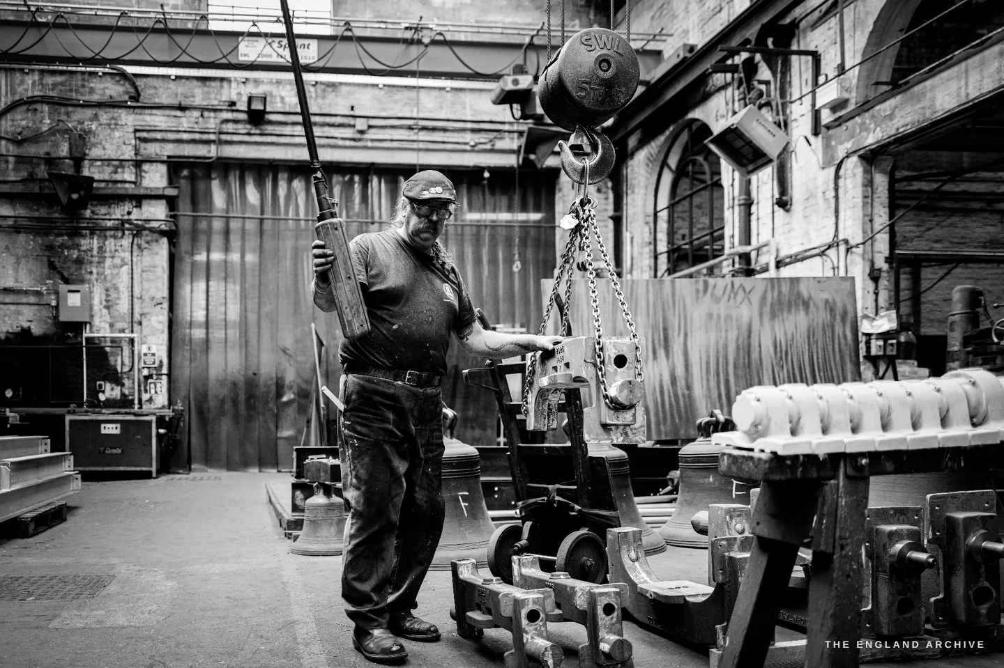 Wide view of Bill standing among ranked bell castings and headstocks in the tall foundry hall, a long steel bar resting on his shoulder, a load gauge hanging on a chain to his right.