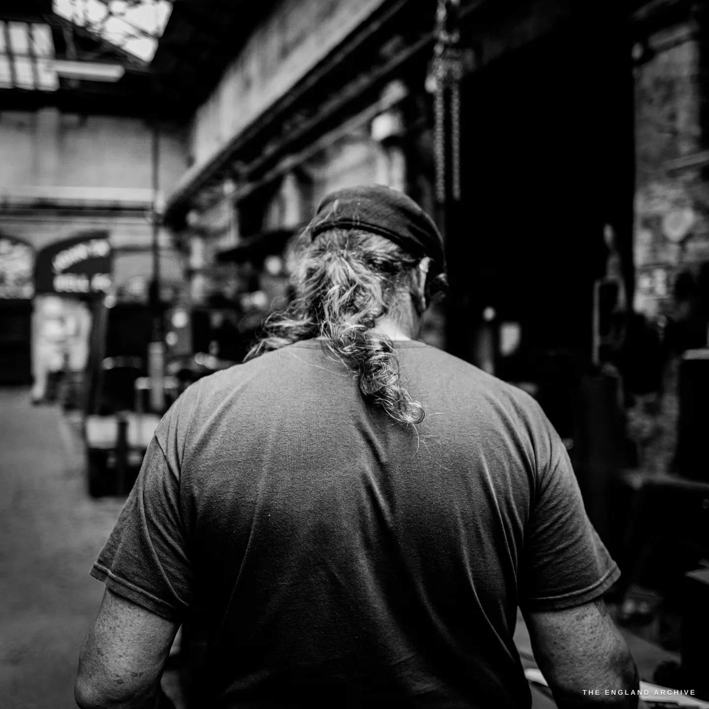 Bill seen from behind, his long grey ponytail down his back over a grey t-shirt, walking away down the foundry aisle into shadow.