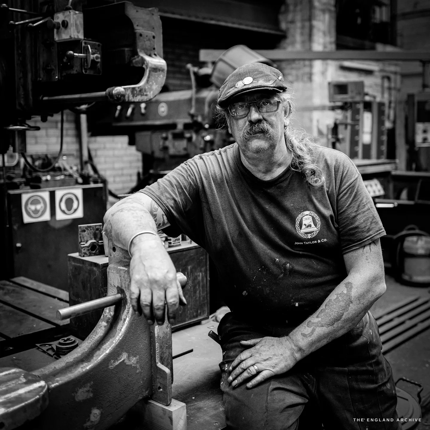 Bill seated and leaning one forearm on a heavy machine, looking straight at the camera, the John Taylor & Co bell mark on his t-shirt, a steel rod in his hand.