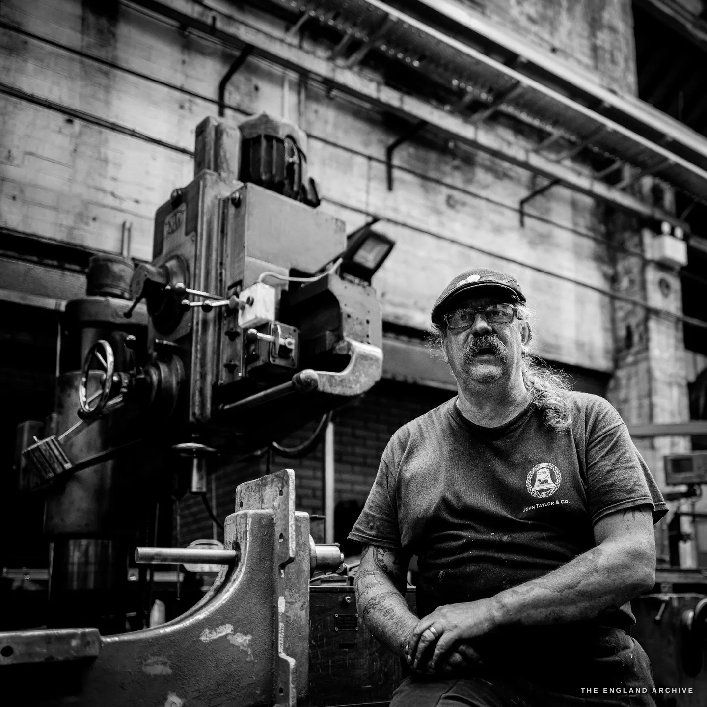 Bill seated against a tall industrial machine and a high concrete wall, looking upward and away, the scale of the foundry rising above him.