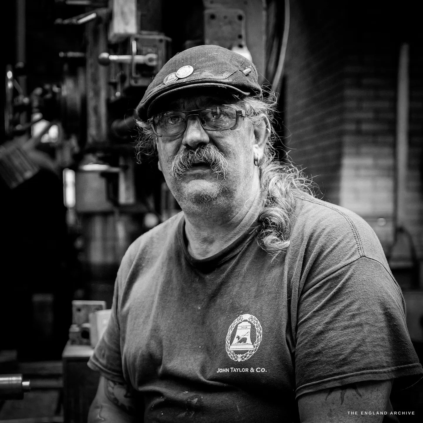 Tight portrait of Bill, the John Taylor & Co bell mark and lettering clear on his t-shirt, the badged cap and glasses lit against the dark machinery behind.