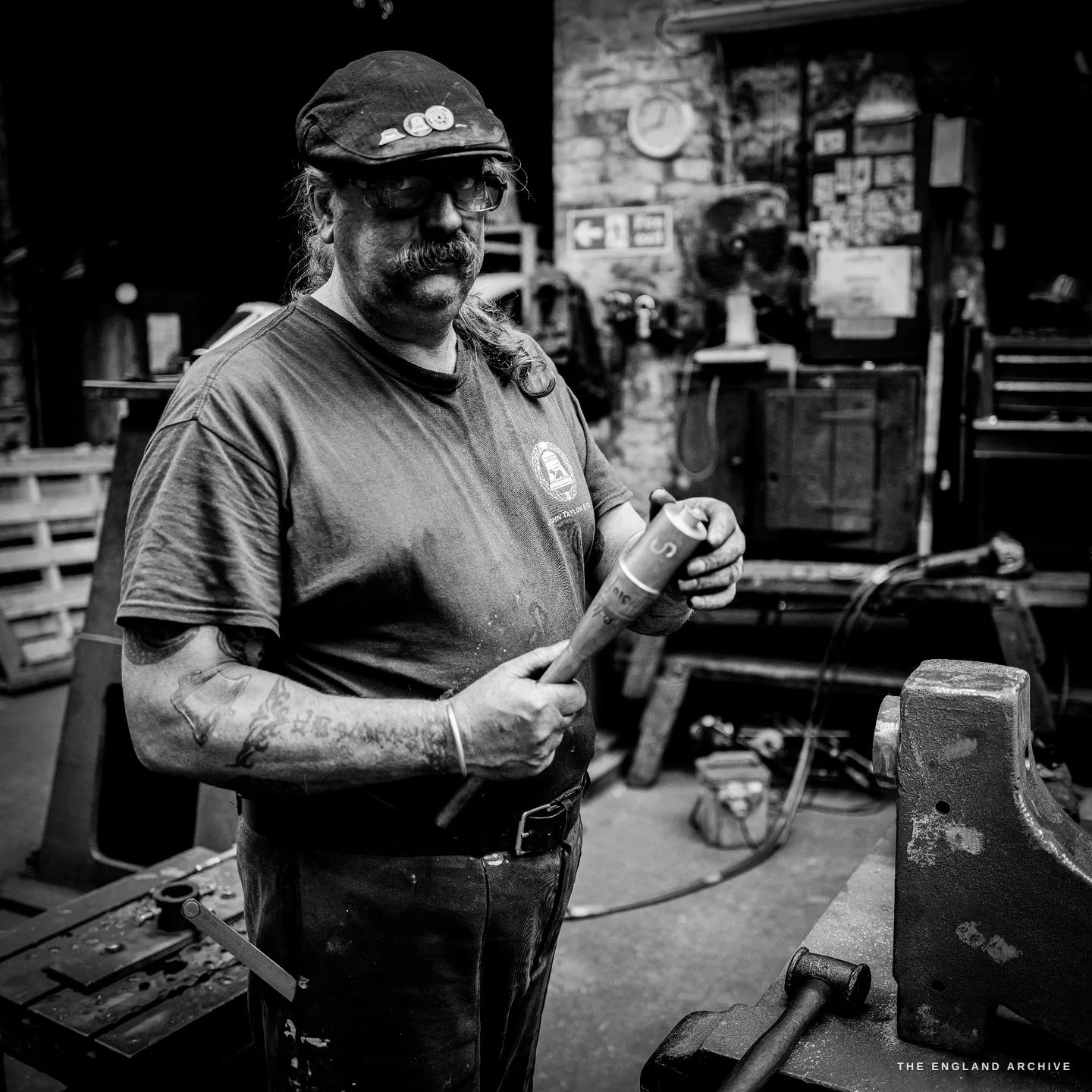 Bill standing at his bench holding a turned metal fitting on a wooden handle, examining it, a brick wall hung with clippings and a fan behind him.