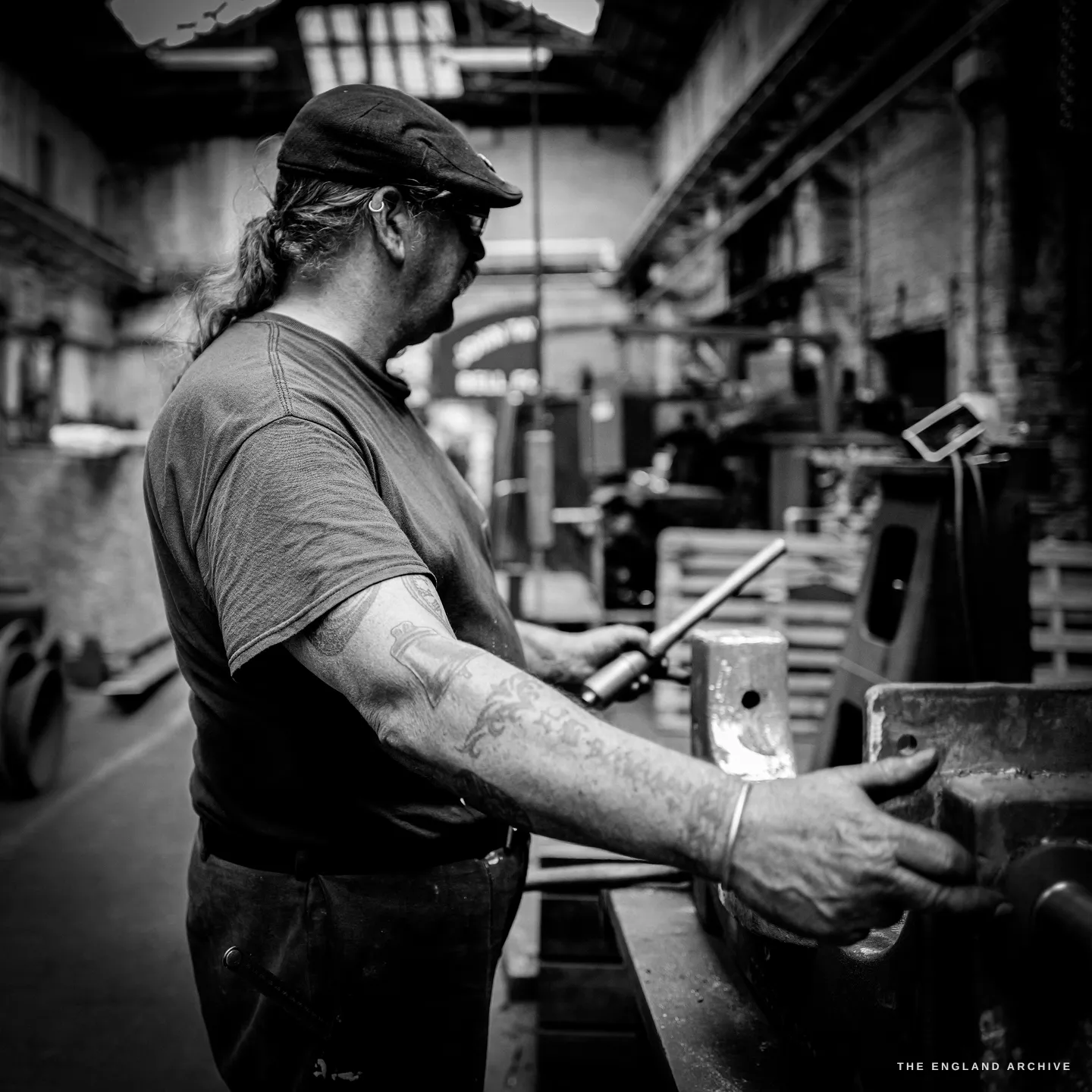 Bill in profile at the vice, both hands on a steel bar laid across a heavy casting, tattooed forearms, the depth of the foundry behind him.