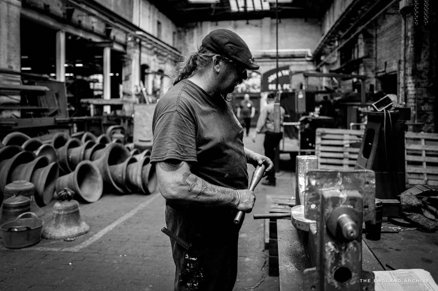 Bill working at a bench in profile, holding a steel bar to a fitting, rows of newly cast bells lining the floor behind him in the foundry hall.