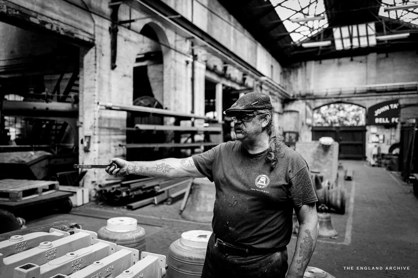 Bill pointing across a row of white cast headstocks with a thin steel rod, the words BELL FOUNDRY visible on an arched sign in the far end of the hall behind him.