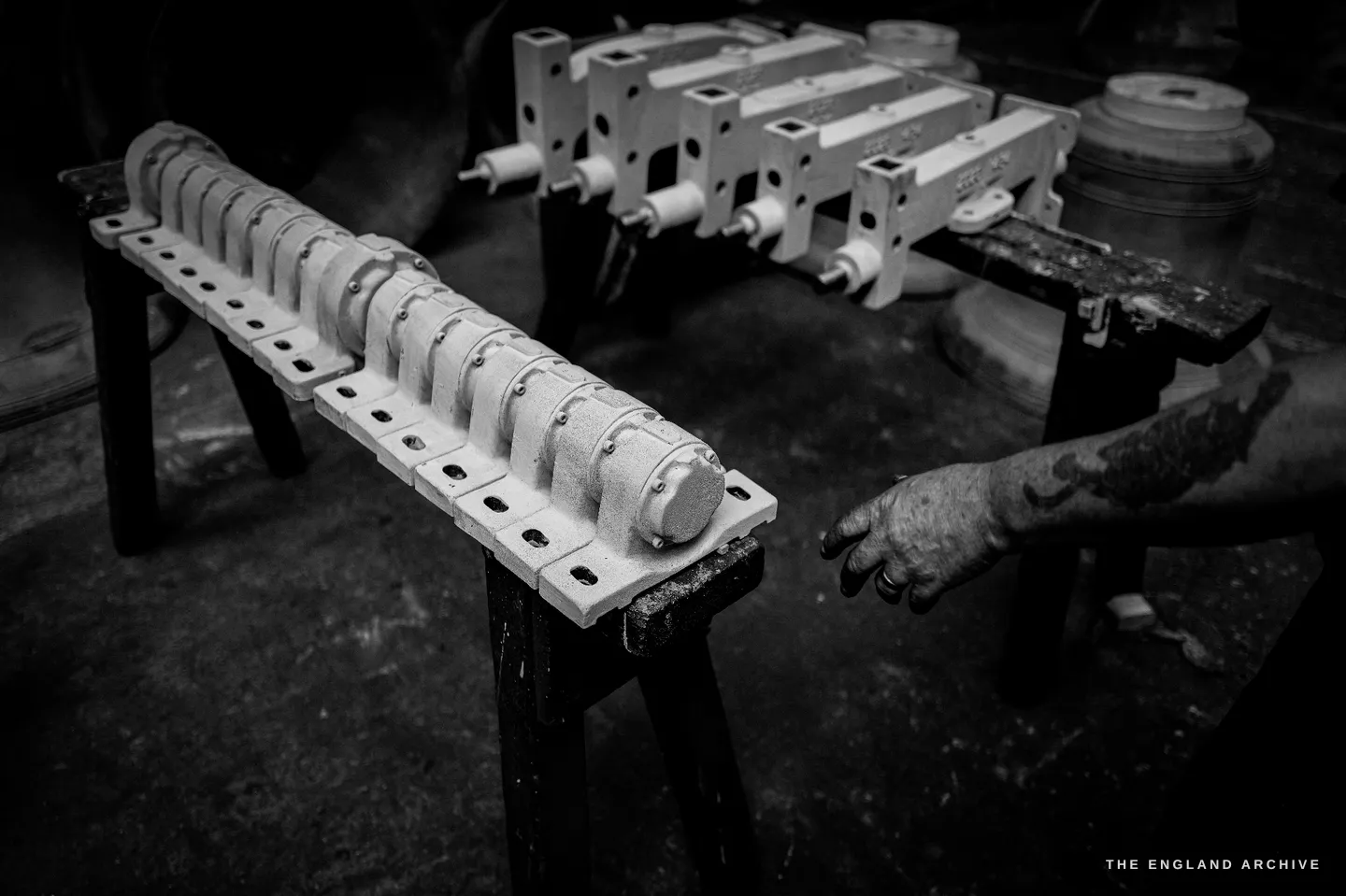 Two sets of pale cast-iron headstock components laid out on trestles, drilled with rows of holes and fitted with white bearing ends, Bill’s tattooed hand reaching in from the right.