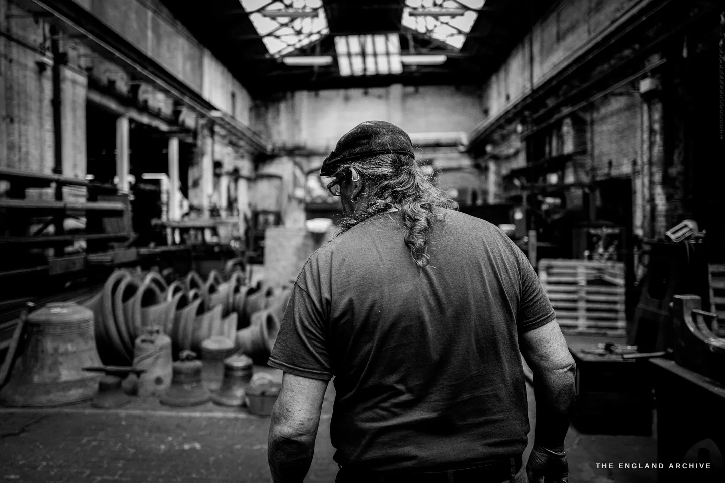 Bill walking away from the camera into the depth of the foundry hall, bells on the floor to his left, the great glazed roof lantern throwing light down the length of the building.