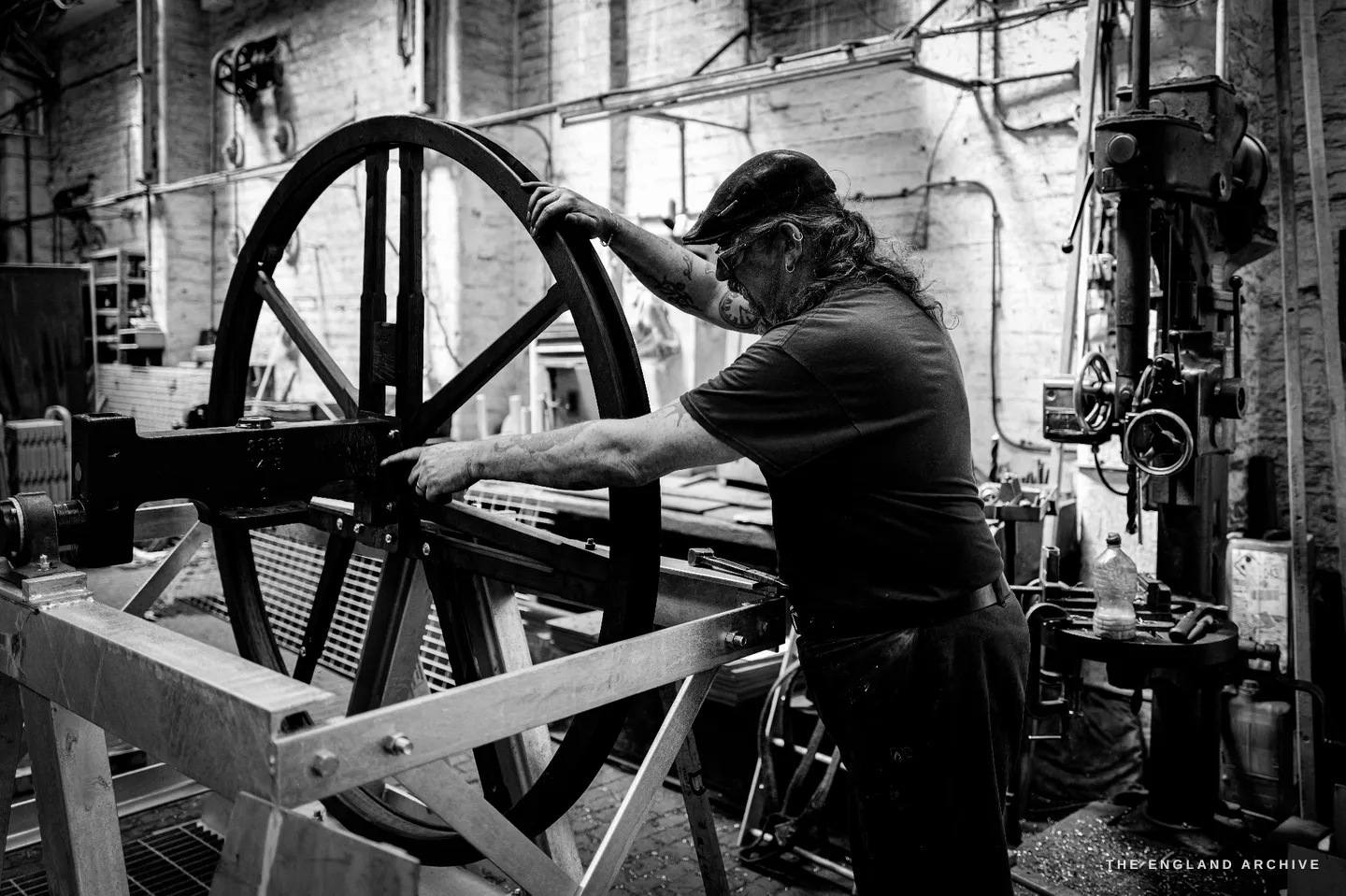 Bill leaning over a large spoked wheel laid flat on a frame, one hand on its rim and the other reaching into the centre, a pillar drill behind him.
