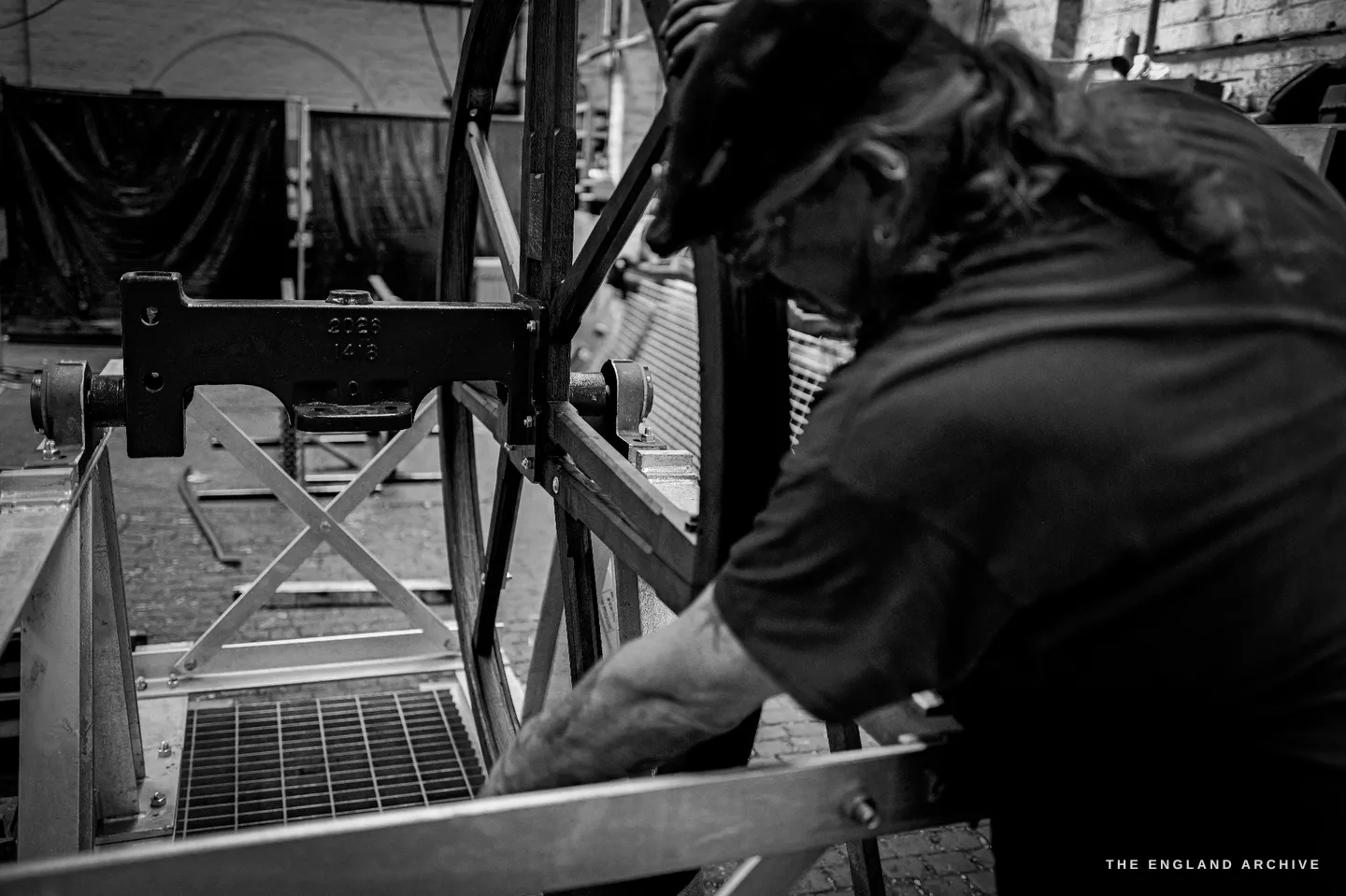 Over Bill’s shoulder as he works on the spoked wheel, a cast number plate reading a foundry job number bolted to the frame in front of him.