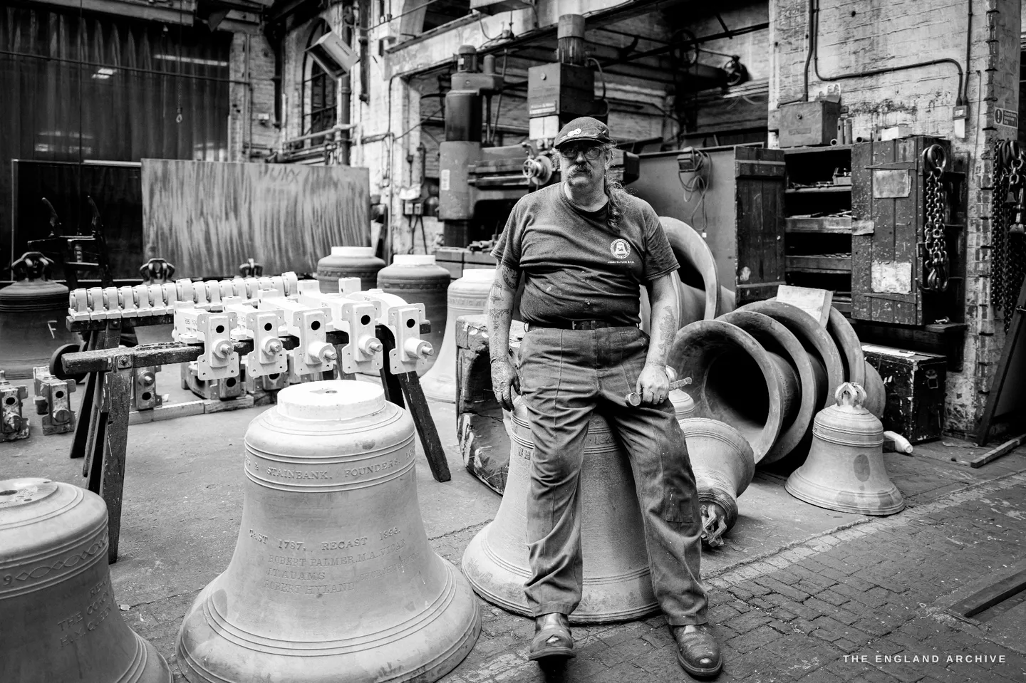 Bill seated on the edge of a large inscribed bell, surrounded on all sides by finished bells of every size, ranks of headstocks behind him in the cluttered yard.
