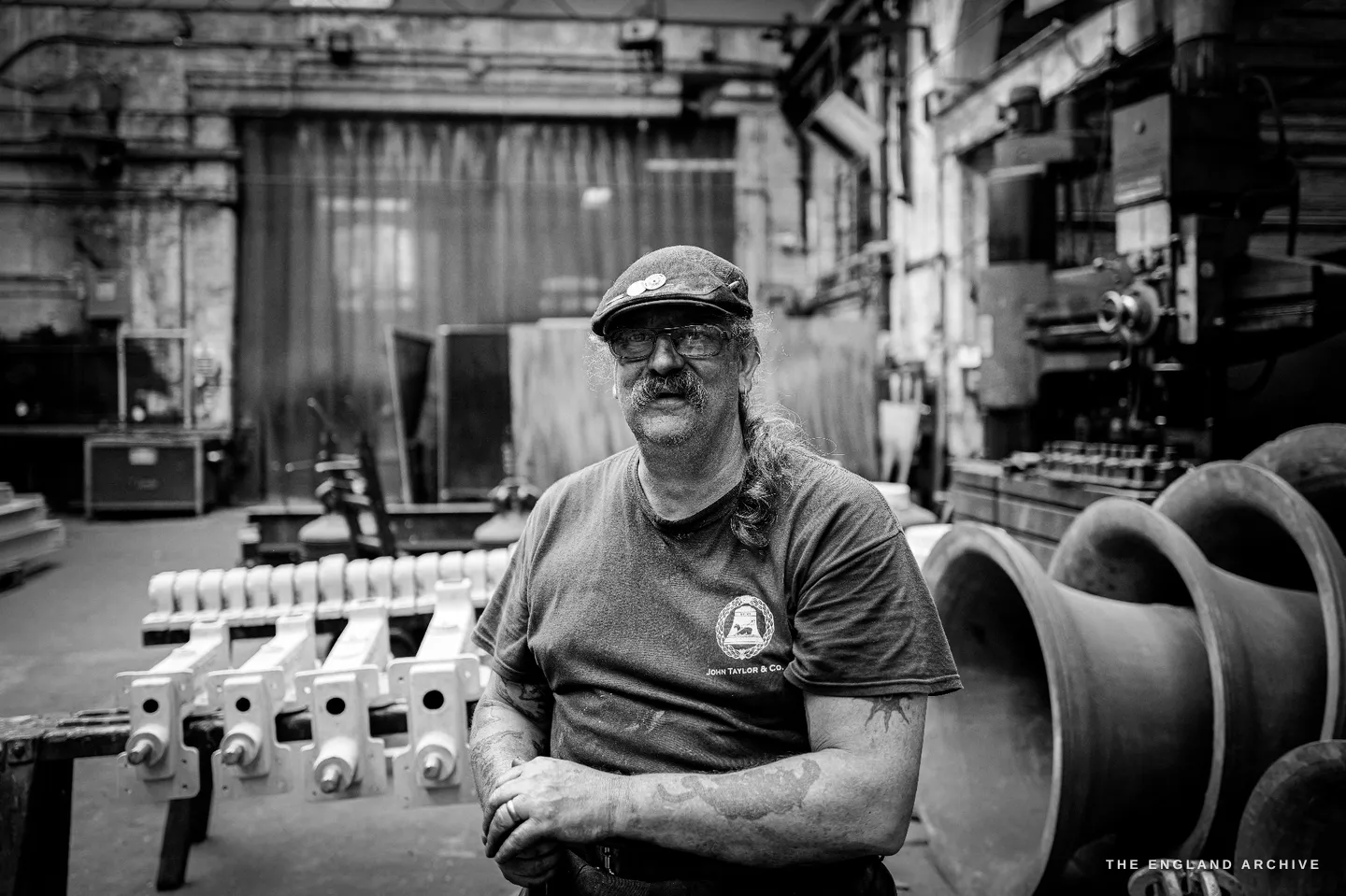 Bill seated in the open foundry hall with white headstocks ranked to his left and a large bell to his right, looking calmly toward the camera.