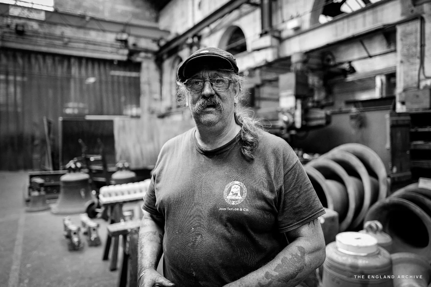 Bill standing square to the camera in the foundry hall, bells and headstocks ranged on either side of him, the great arched roof lantern above.