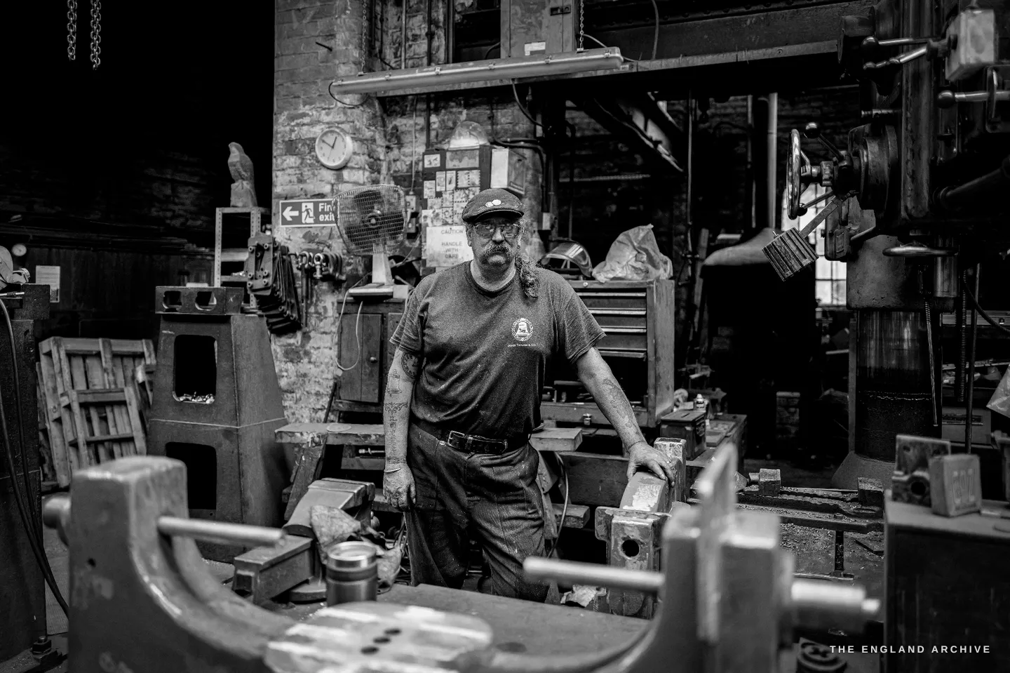 Bill standing at his workbench beside a brick pillar pinned with clippings and an old electric fan, a heavy vice clamped in the foreground.