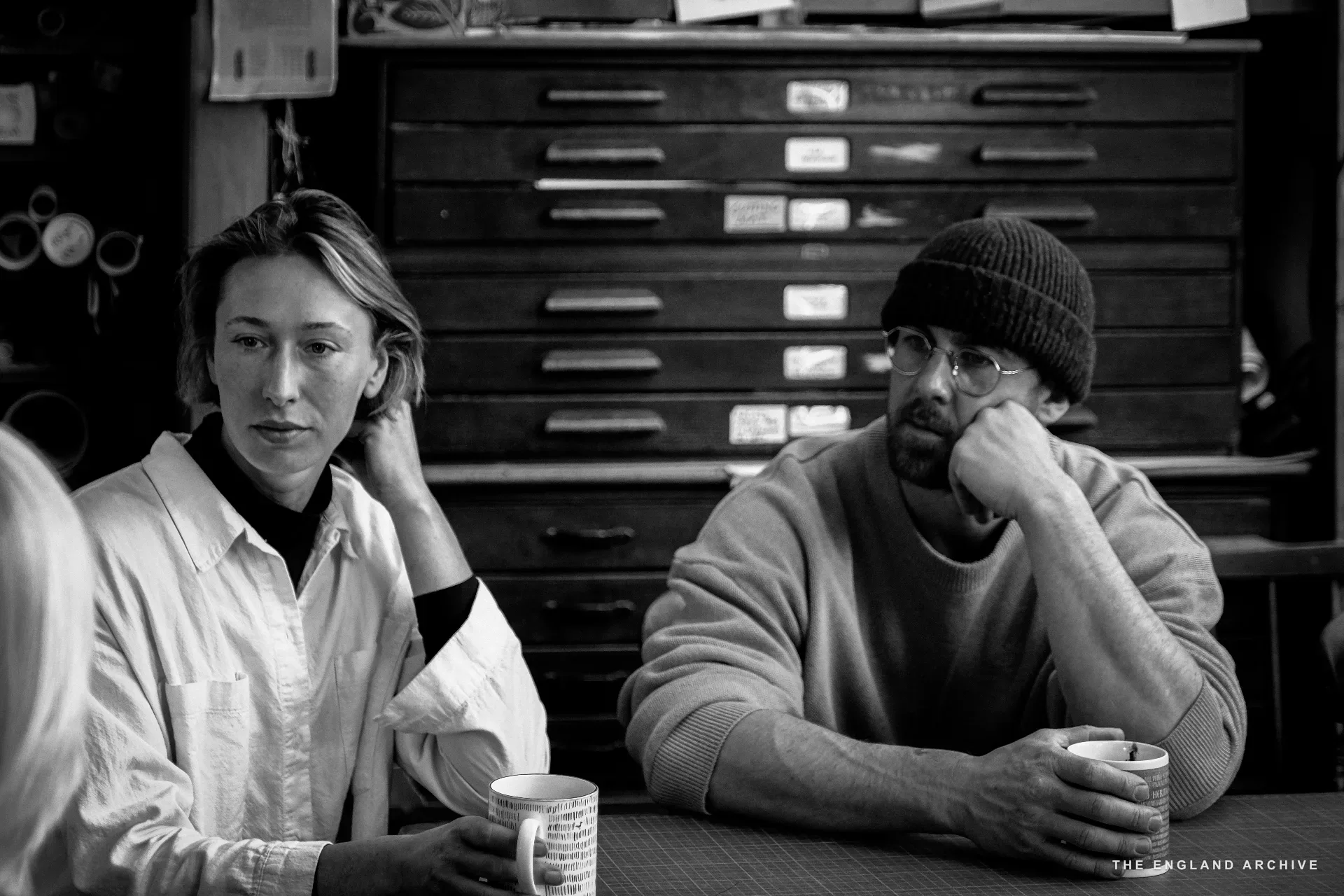 Two of the workshop team at the front-room table - a young woman in a white shirt on the left, a man in a dark beanie and round glasses on the right - both holding mugs of tea. A printer's tray cabinet stands behind them.