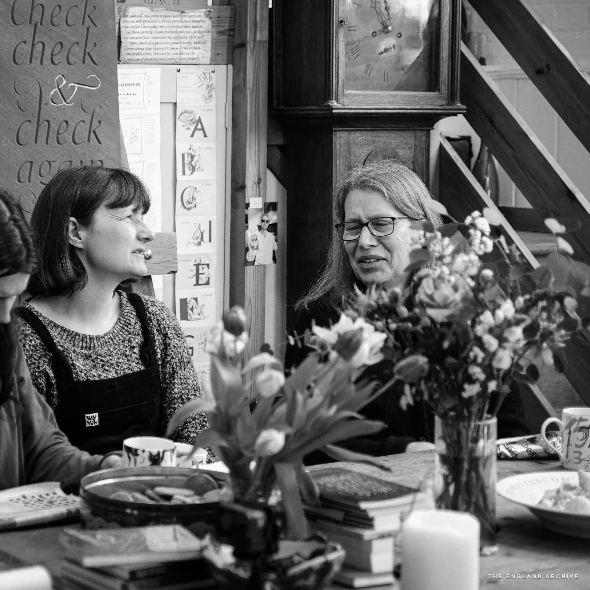 The big table in the front room. A young woman with dark hair on the left in profile, Lida Kindersley on the right in glasses, looking past the table; tulips in a vase between them, a longcase clock and a slate sign reading 'check check & check again' on the wall behind.