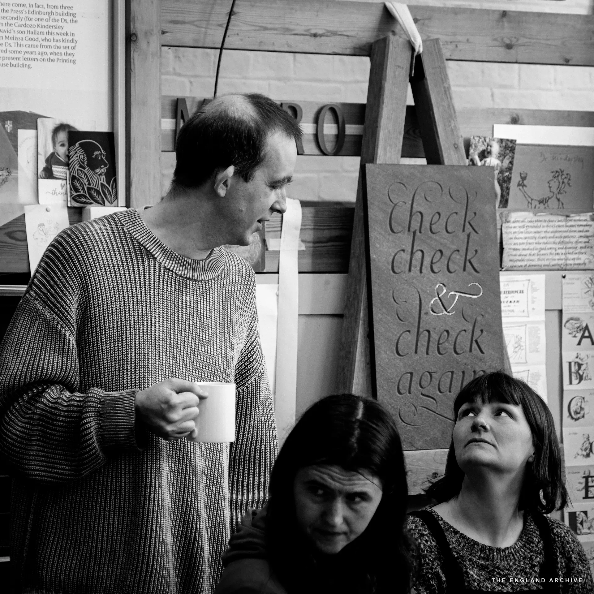 A workshop visitor in a knitted jumper standing beside the front-room table holding a mug, mid-sentence, with two seated women looking up at him. Behind him stands the slate sign reading 'check check & check again' and the workshop wall lettering reads 'MORO'.