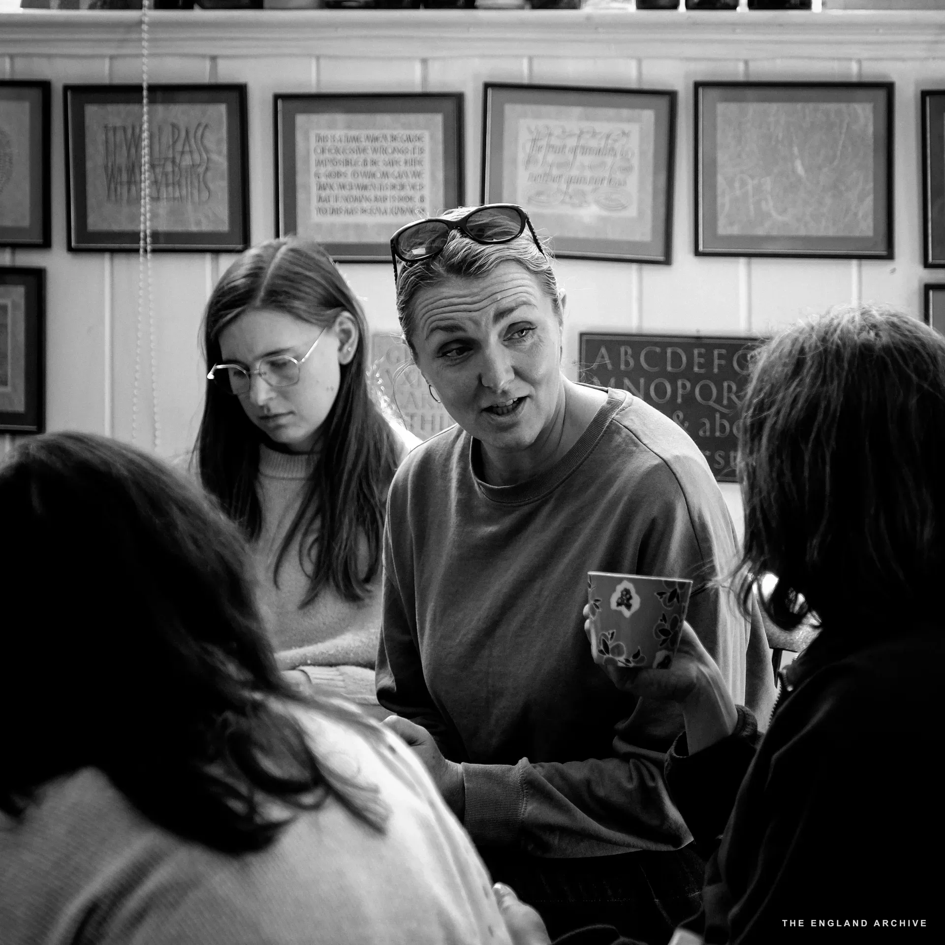 Three figures in conversation around the front-room table. Lida Kindersley in the centre with sunglasses pushed up onto her head, holding a floral mug, talking. A young woman with thin glasses to the left listens. The wall behind is hung with framed letter samples and an alphabet panel.