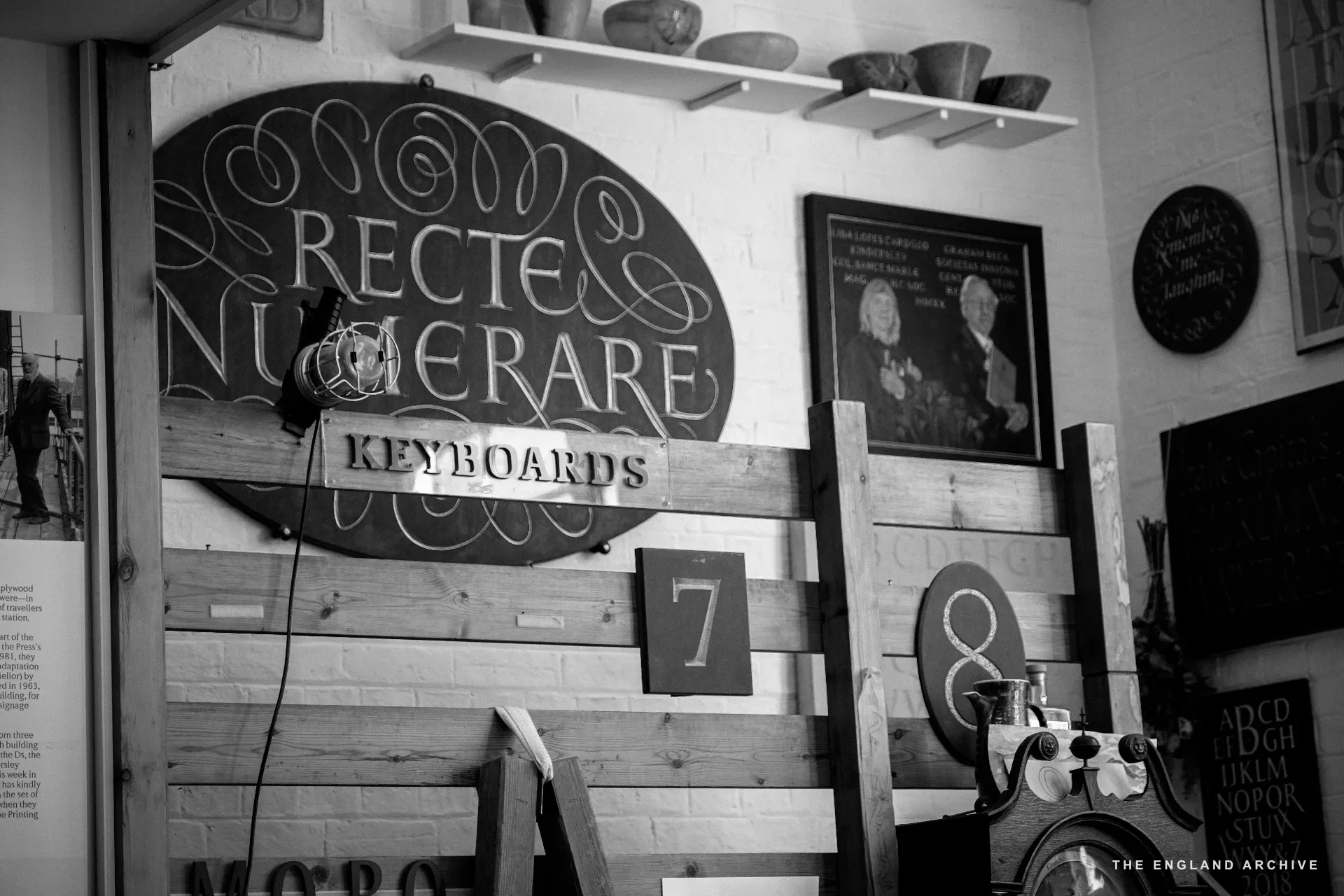 The workshop's carved wooden 'RECTE NUMERARE KEYBOARDS' sign mounted above stacked pallets and bowls on a shelf. A pendant lamp drops in from above; the workshop's alphabet samples are just visible to the right.