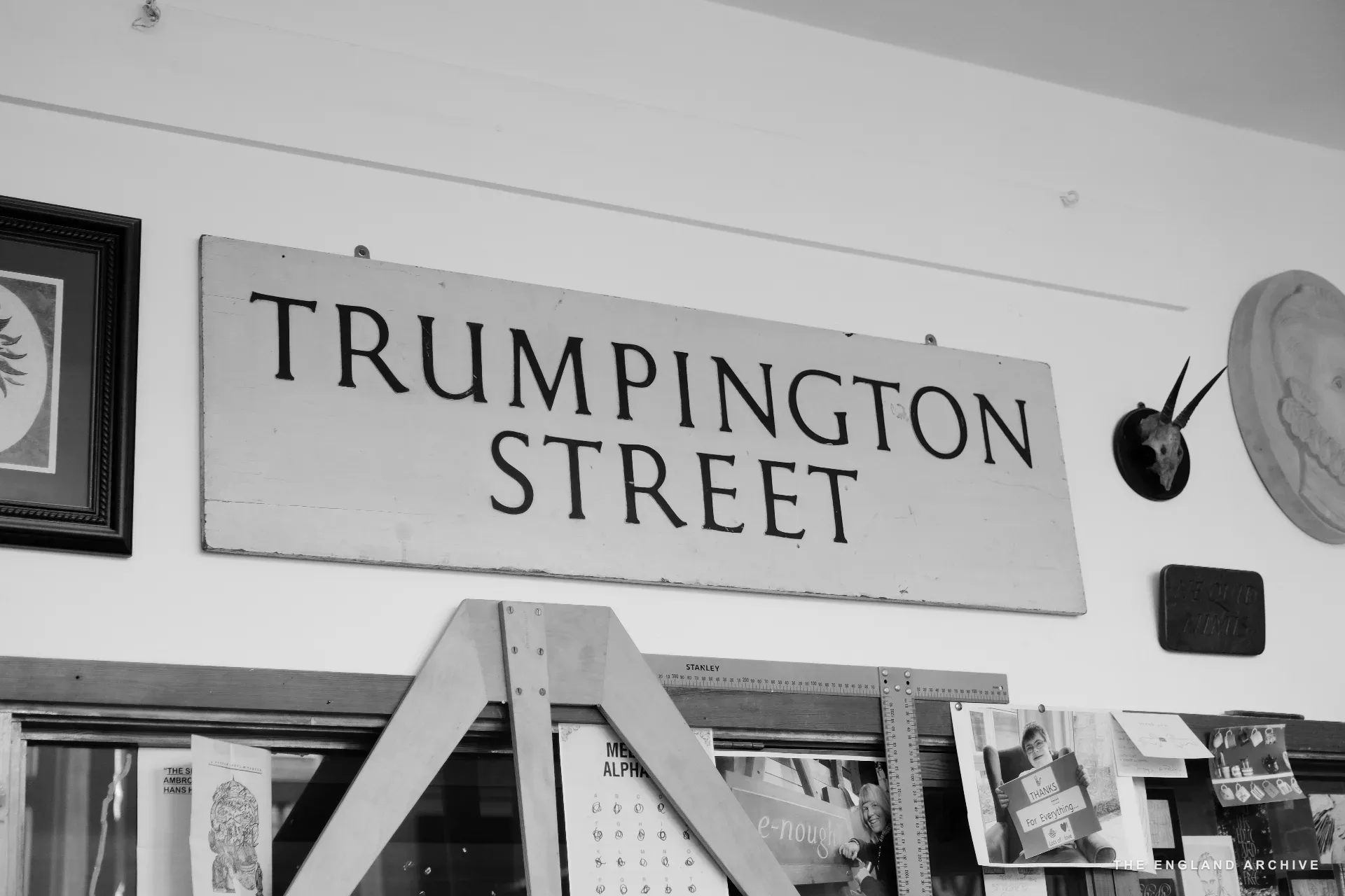 A salvaged painted street sign reading 'TRUMPINGTON STREET' hung high on a workshop wall, with a Stanley combination square ruler propped on the easel below and pinned-up reference photographs and lettering samples around it.