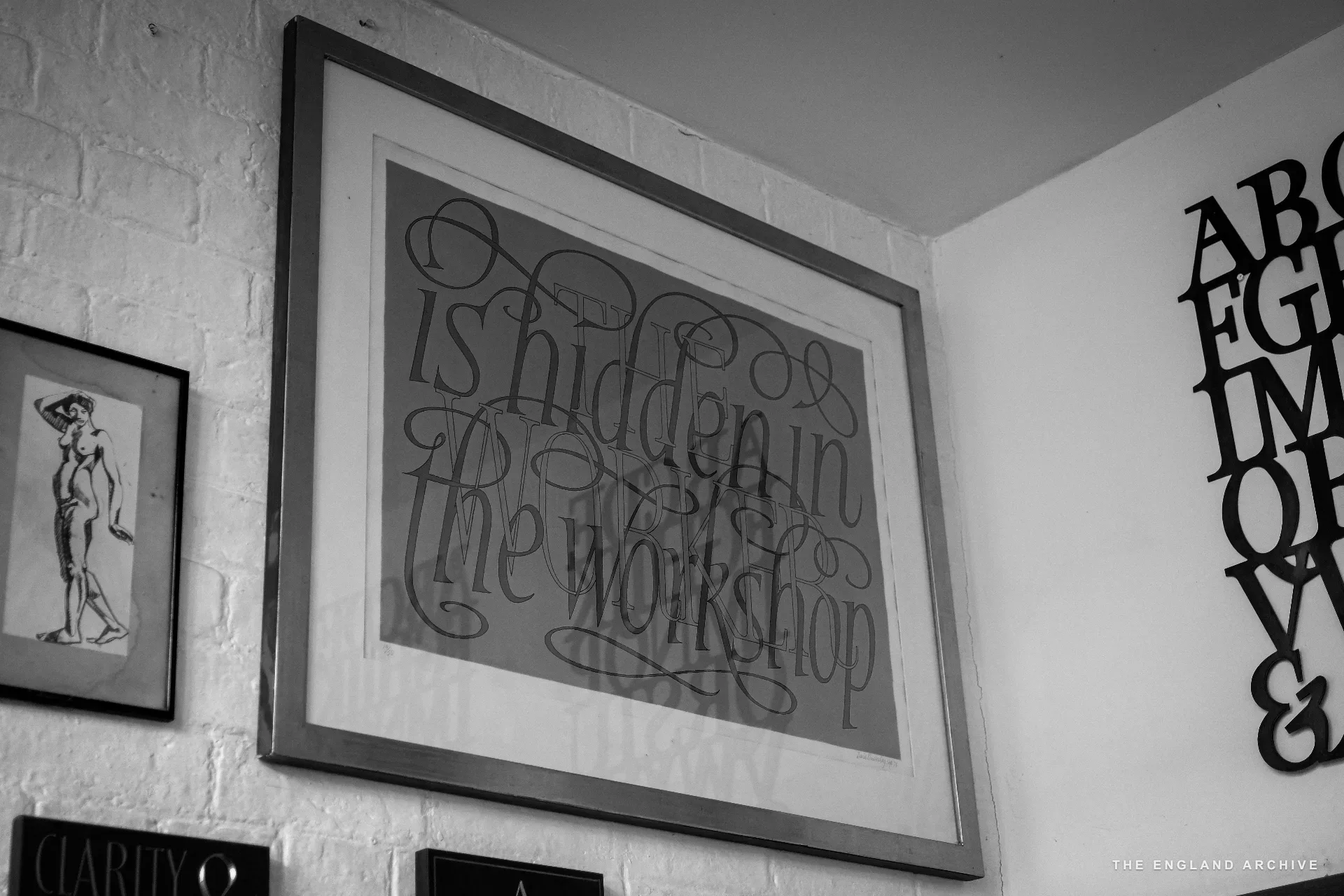 A large framed letter-cut piece on the workshop wall reading 'is hidden in the workshop' in flowing italic capitals. A smaller framed nude figure-study hangs alongside it on the brick wall.