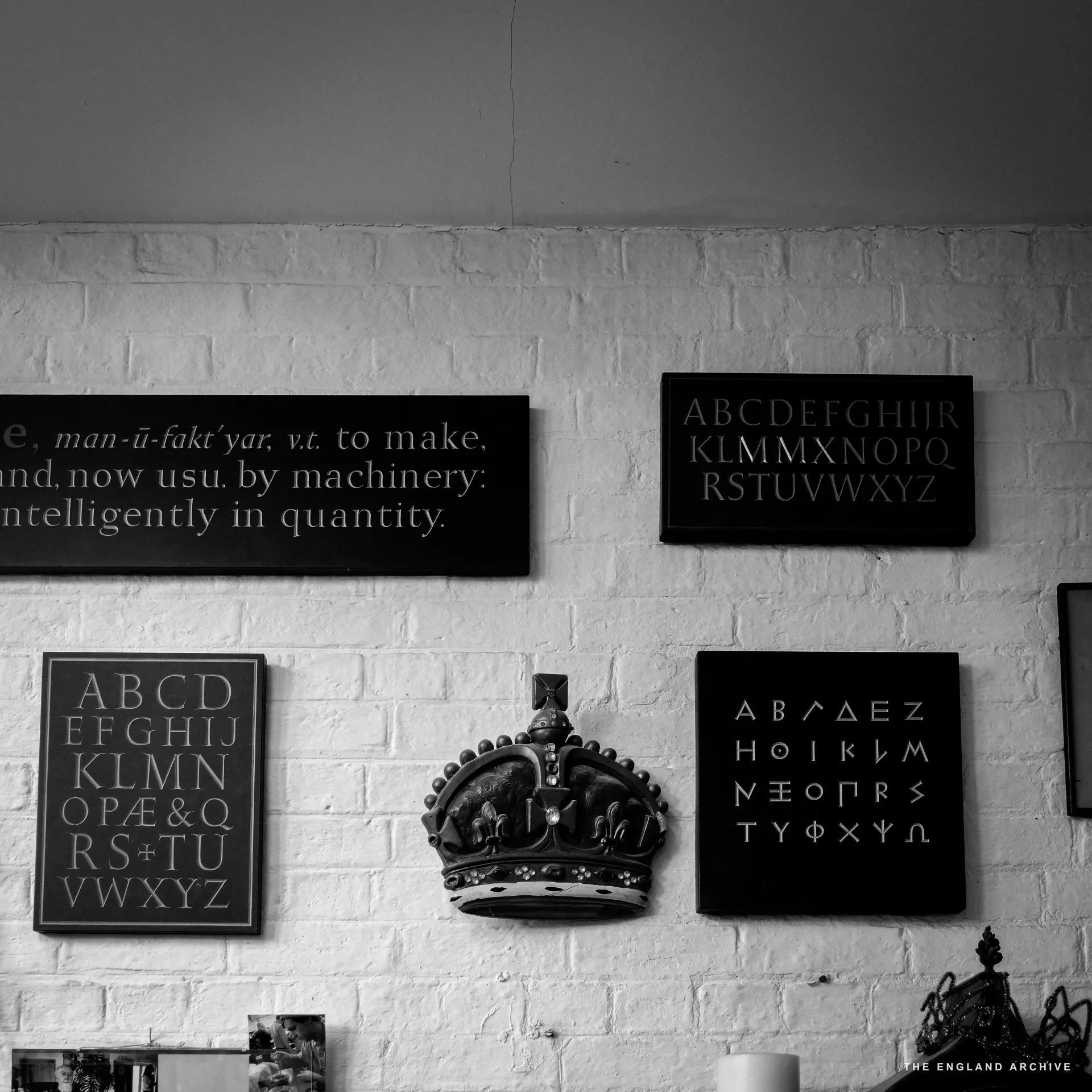 A wall display of letter-cut samples mounted on white-painted brick: a slate sign reading 'man-u-fakt'yar, v.t. to make, and now usu. by machinery: intelligently in quantity', alphabet panels in roman and Greek, and a wooden carved heraldic crown.