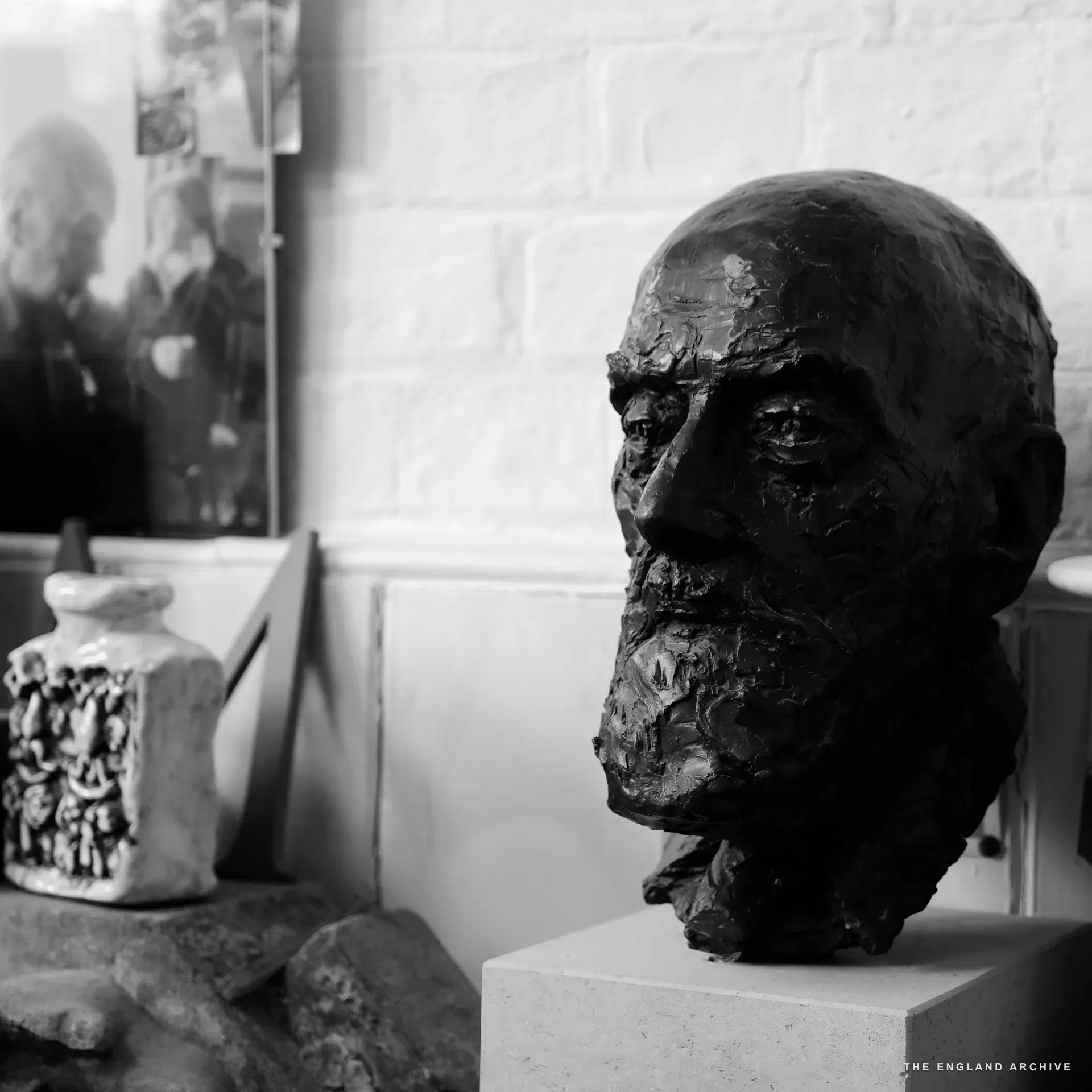 A bronze portrait head on a stone plinth in the workshop, lit from the side; in the soft-focus background a mirror reflects two figures. A small ceramic vessel and a stone offcut sit beside the plinth.