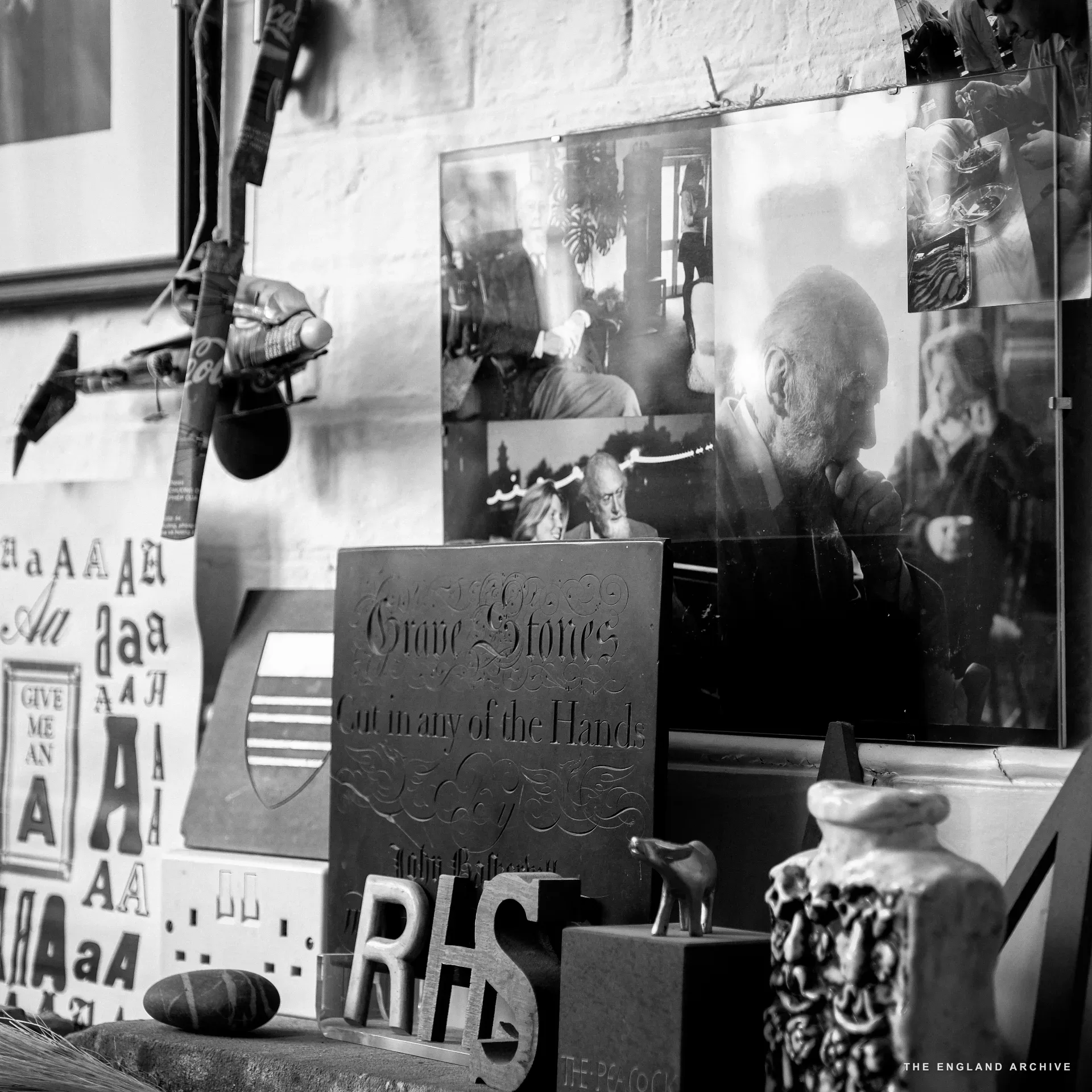 Workshop corner with a slate plaque reading 'Grave Stones / Cut in any of the Hand' propped against the wall, large carved letters 'RHS' in front of it, and on the wall above a black and white photograph of an older bearded man at work.