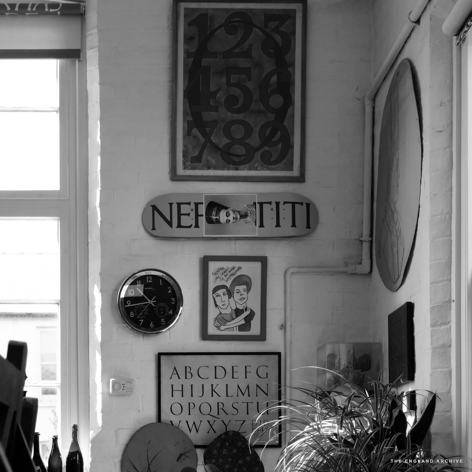 The workshop's window corner: a framed numerals sample reading '1234567890' hangs centre, a skateboard-deck mounted with cut letters reading 'NEFERTITI' beneath, a round wall clock, an ABCDEF alphabet tile and a potted plant filling the corner.