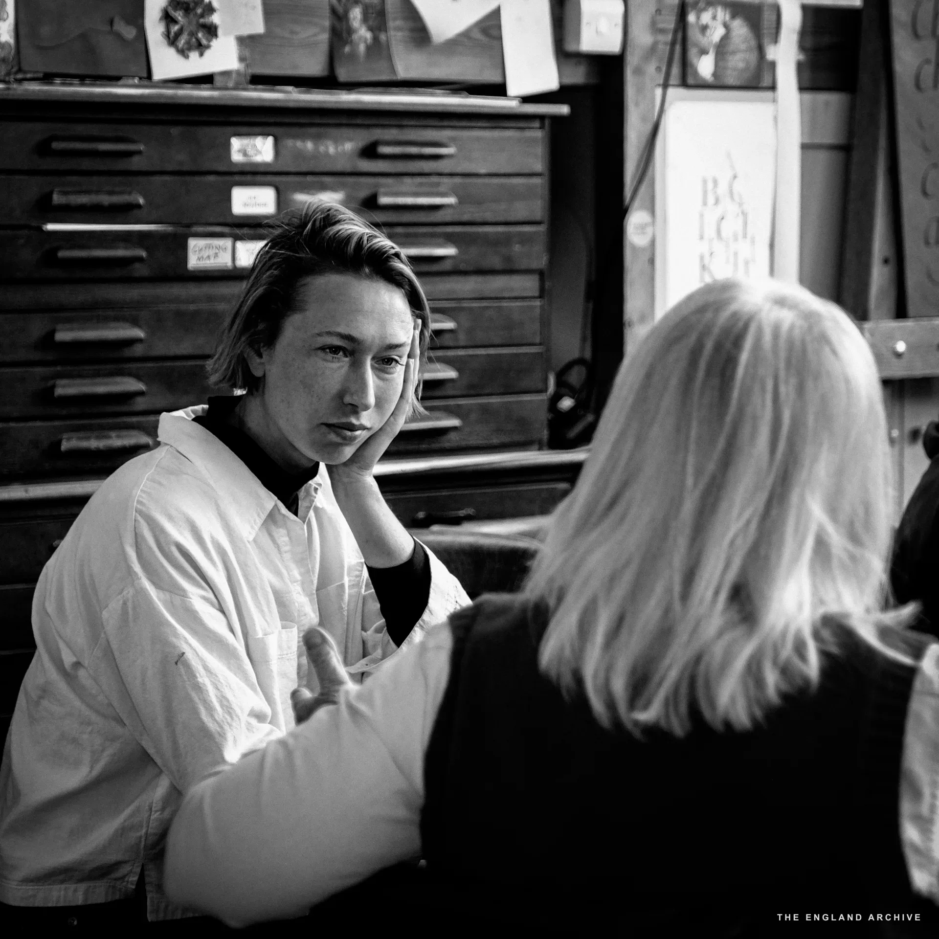 A young workshop member with shoulder-length hair in a white shirt, hand resting against face, mid-conversation with a blonde-haired older woman whose back is to the camera. A printer's tray cabinet stands behind, its drawers labelled.