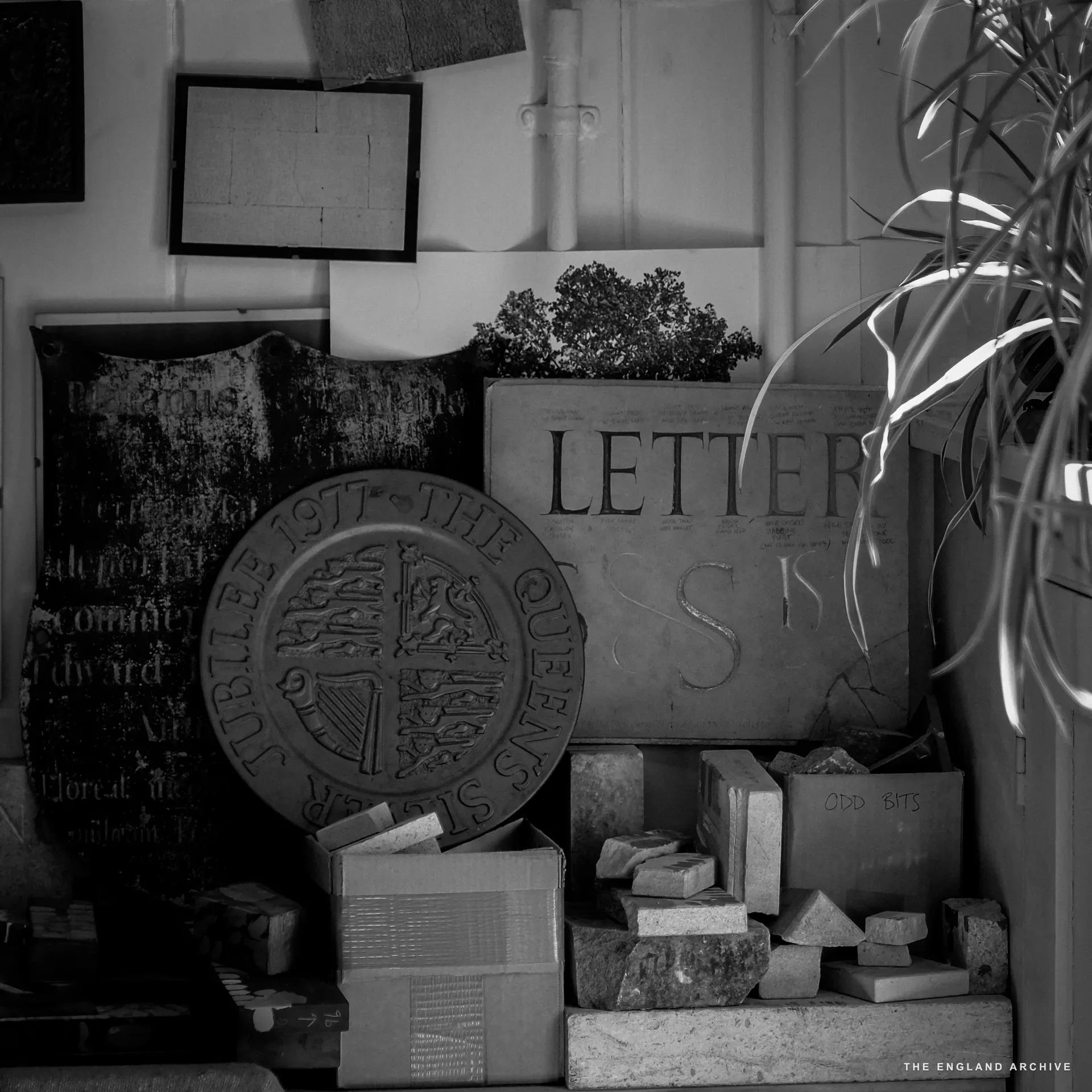 A workshop corner stacked with finished and offcut stone: a Queen's Silver Jubilee 1977 carved stone roundel on the floor, a slate panel reading 'LETTER S' propped behind it, boxes of cut blocks and offcuts on a low shelf, a tall plant to one side.