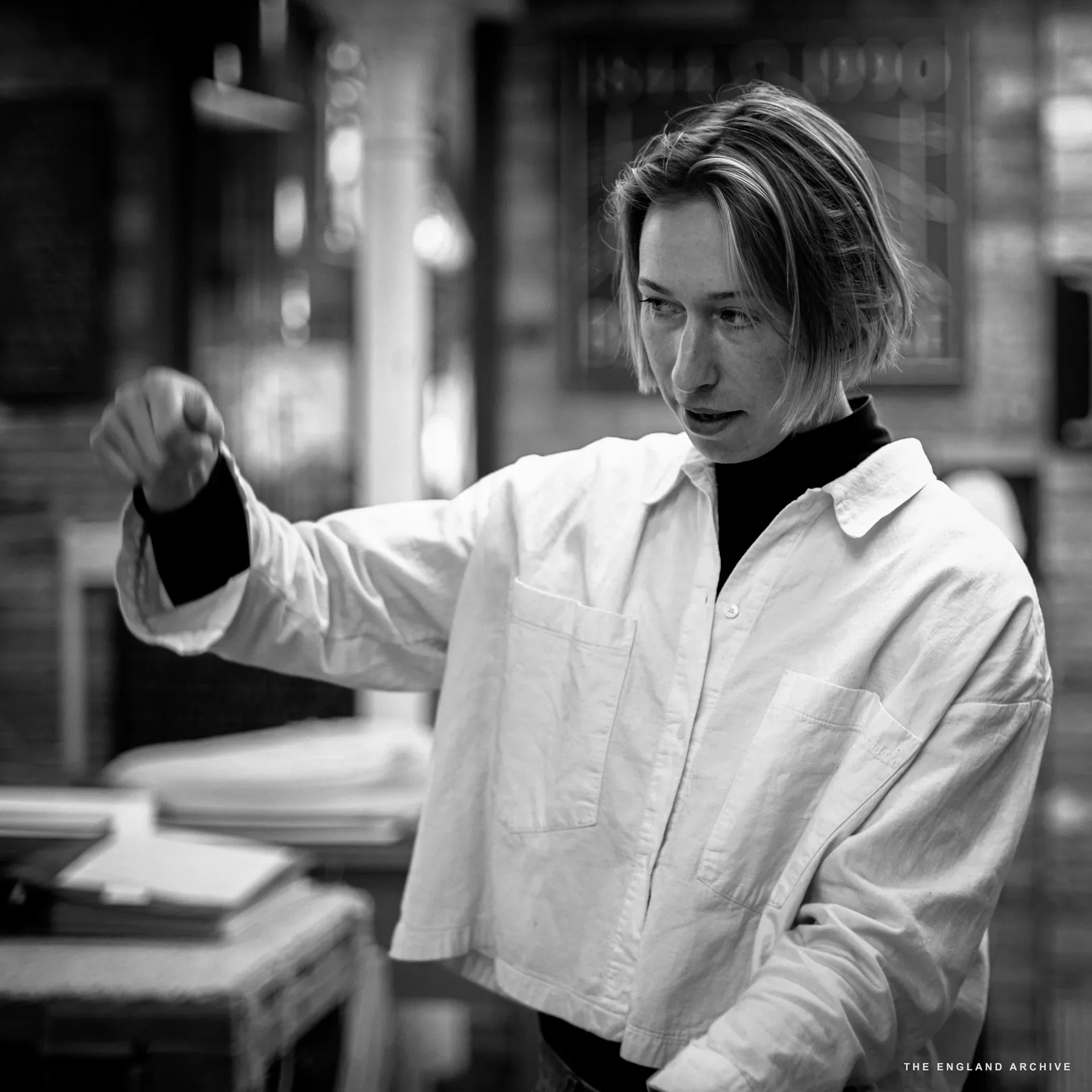 Roxanne Kindersley standing in a white shirt over a black turtleneck, hand raised mid-gesture as she explains. Behind her, a workshop window etched with the dates '1884-1990' is just visible.
