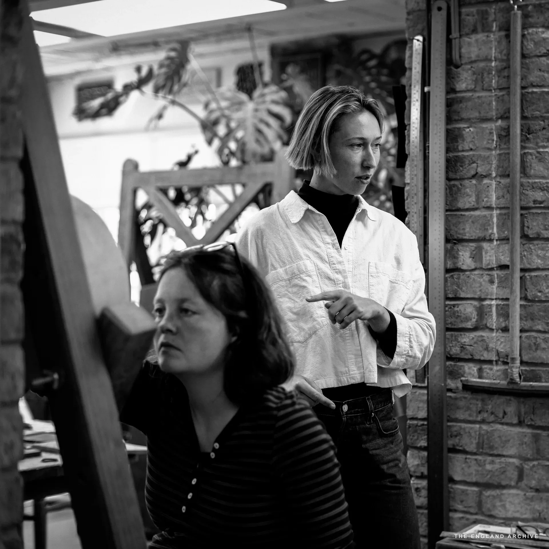 Roxanne Kindersley standing in a white shirt, hand raised pointing at something out of frame, behind a seated woman in a striped top who looks the same way. The workshop's brick wall and ruler edge frame the right side.