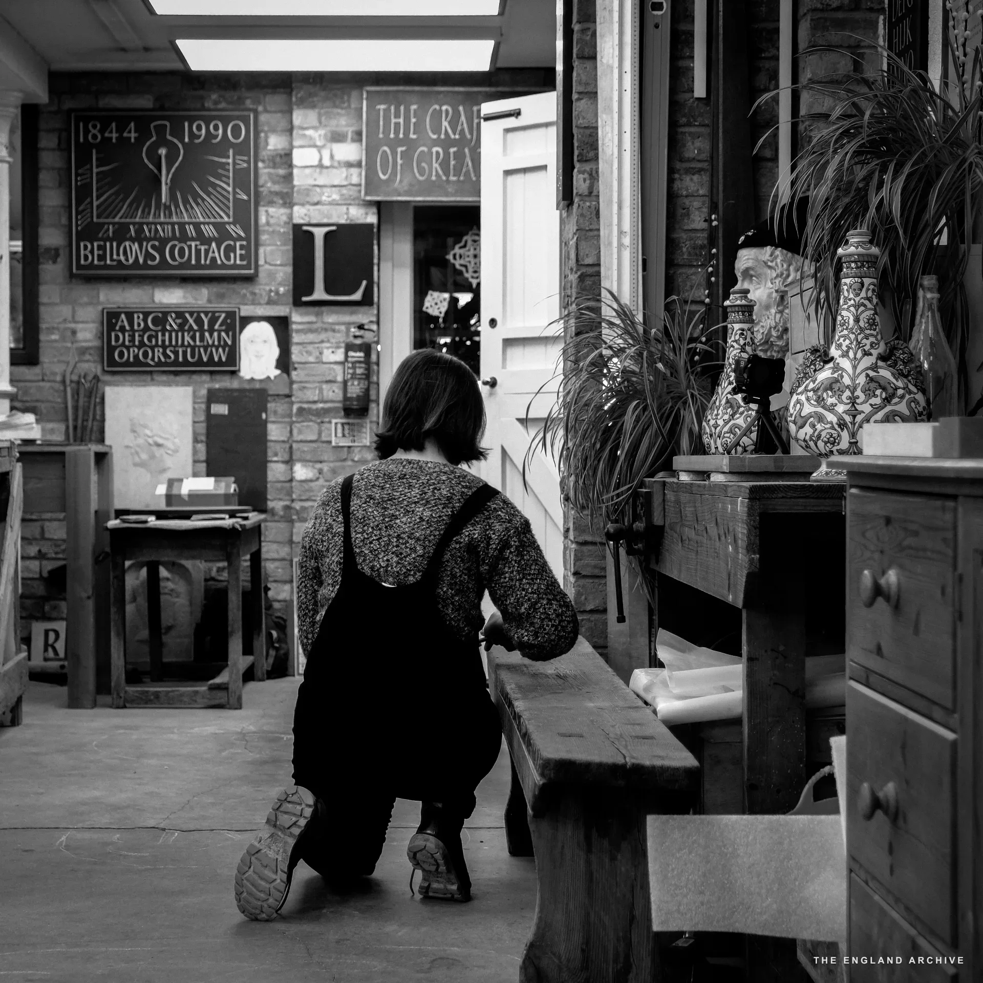 A figure in dungarees crouching by a low wooden bench in the main workshop, light falling through a skylight above. On the wall behind: a slate sign reading 'BELLOWS COTTAGE 1844-1990', alphabet samples, and a slate inscribed 'THE CRAFT OF GREAT...'