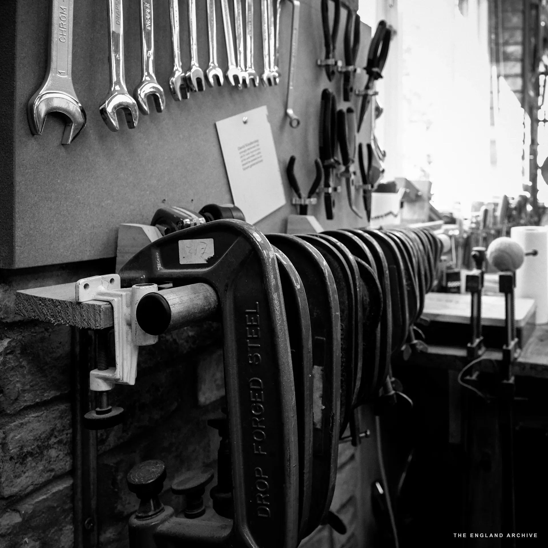 The workshop's tool wall: a row of chrome spanners hung along the top, pliers and wire-cutters below, a large drop-forged steel sash clamp stretched across the lower bench, with chisels and other hand-tools racked behind.