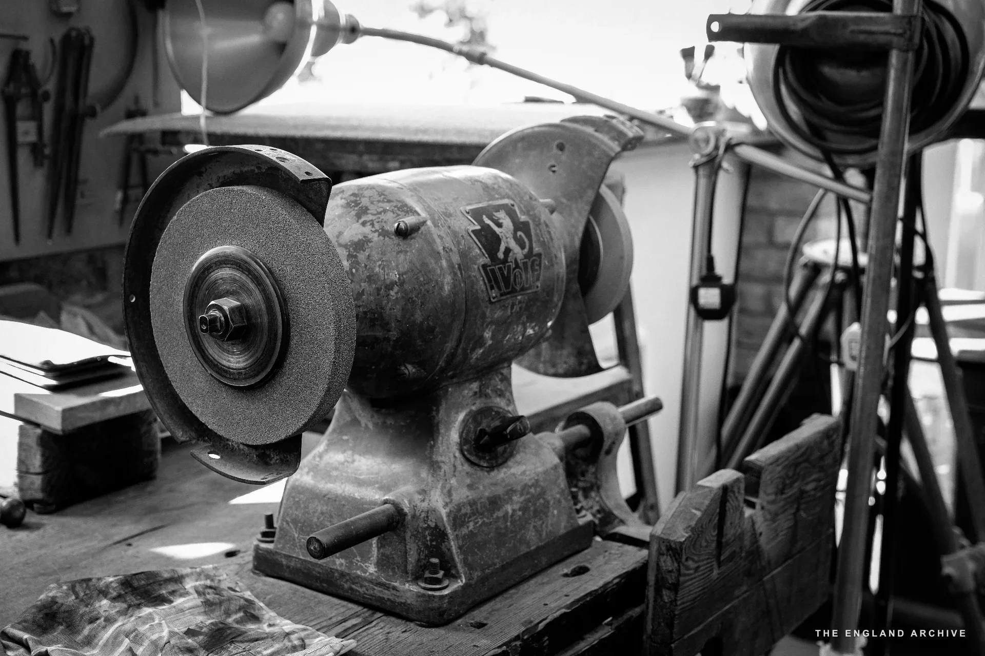 A heavy bench grinder on the workshop's tool bench, twin grinding wheels caked with stone dust, the metal body marked 'X3 / WMP'. Behind it, the workshop's window light.
