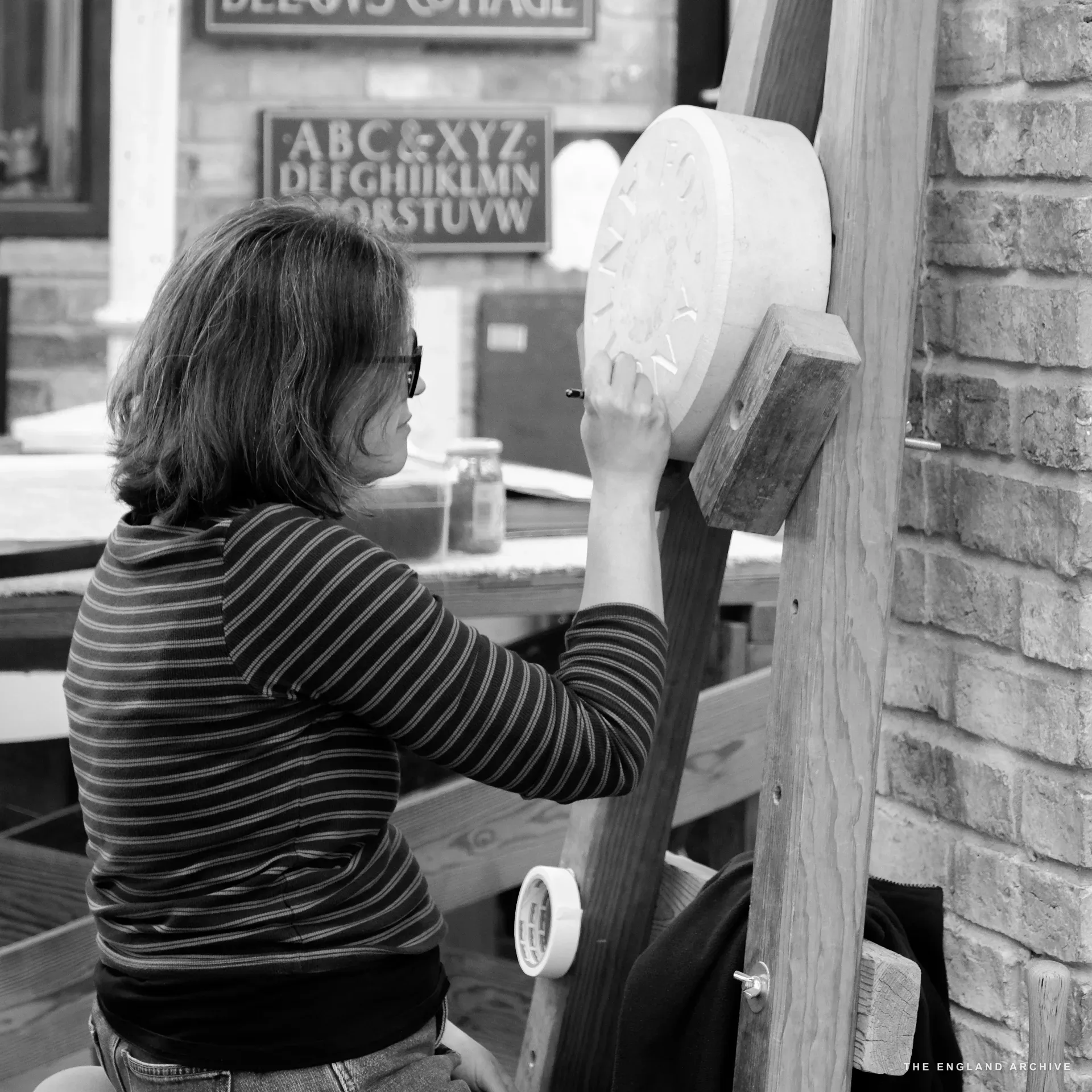A workshop letterer seated at the upright easel, working with a chisel and dummy on a circular stone disc held in a wooden cradle. Behind her, the 'BELLOWS COTTAGE 1844-1990' slate and an alphabet panel are visible on the wall.