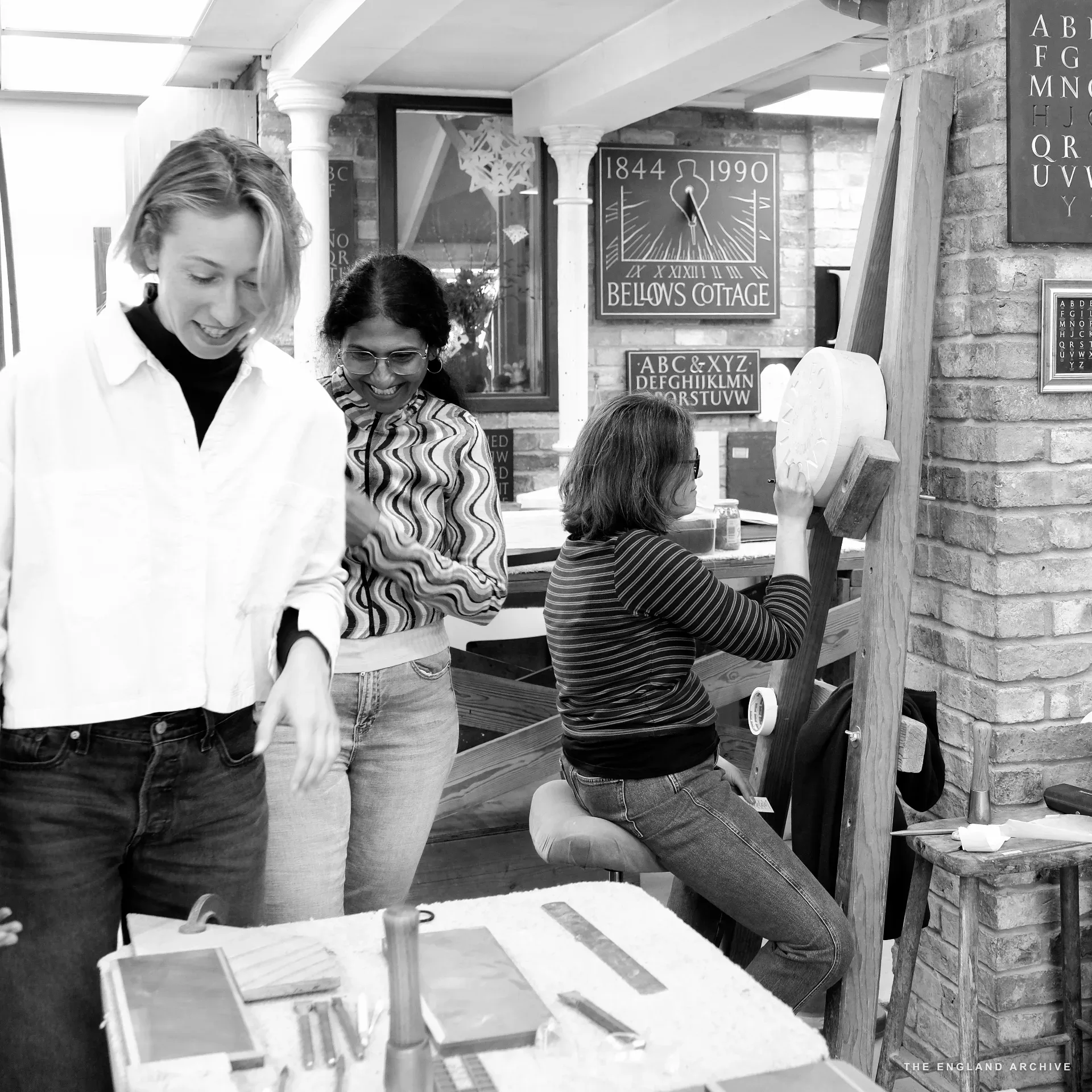 Three workshop members at a bench: Roxanne Kindersley standing left in a white shirt, a young woman in a striped top in the centre, and a letterer on the right in a striped top working with a chisel on a circular stone disc. The alphabet panels are visible on the brick wall.