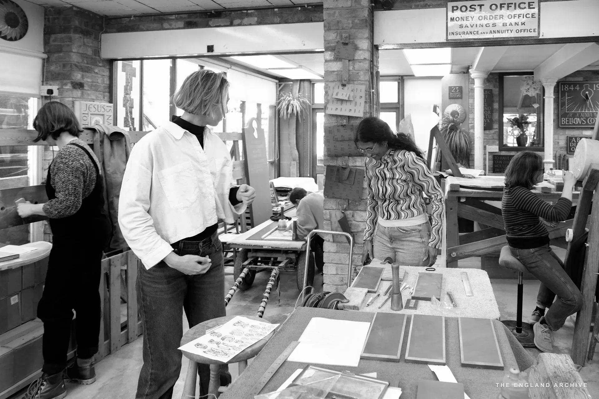A wide view across the workshop's back room: several team members at benches working on different pieces, light streaming in from the back, a salvaged painted 'POST OFFICE / MONEY ORDER OFFICE / SAVINGS BANK' sign hung on the high wall.