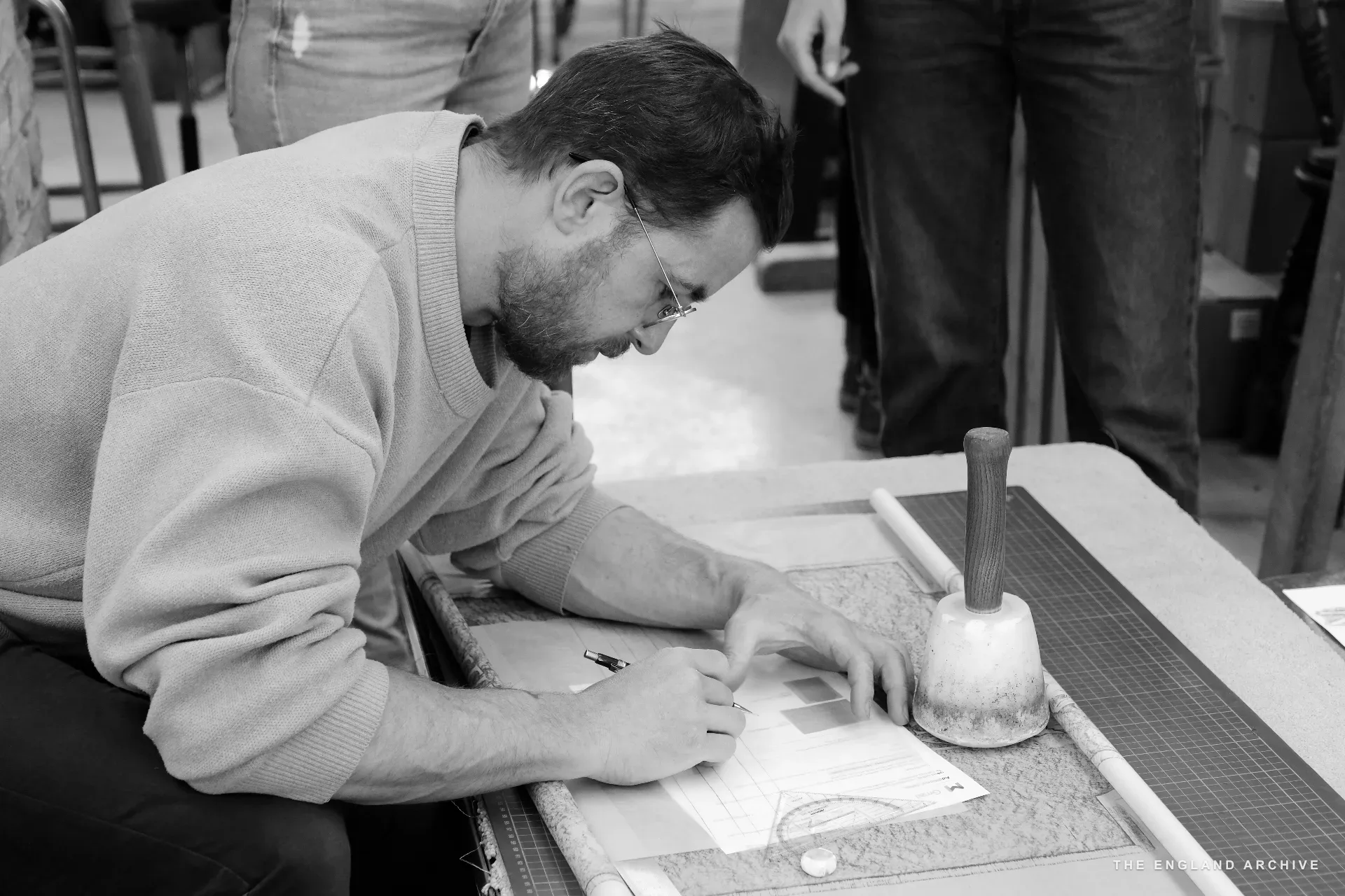 A bearded man in a pale jumper and glasses leaning over a stone laid flat on the bench, marking out lettering with a pencil; a heavy wooden mallet rests beside him on a rubber mat.