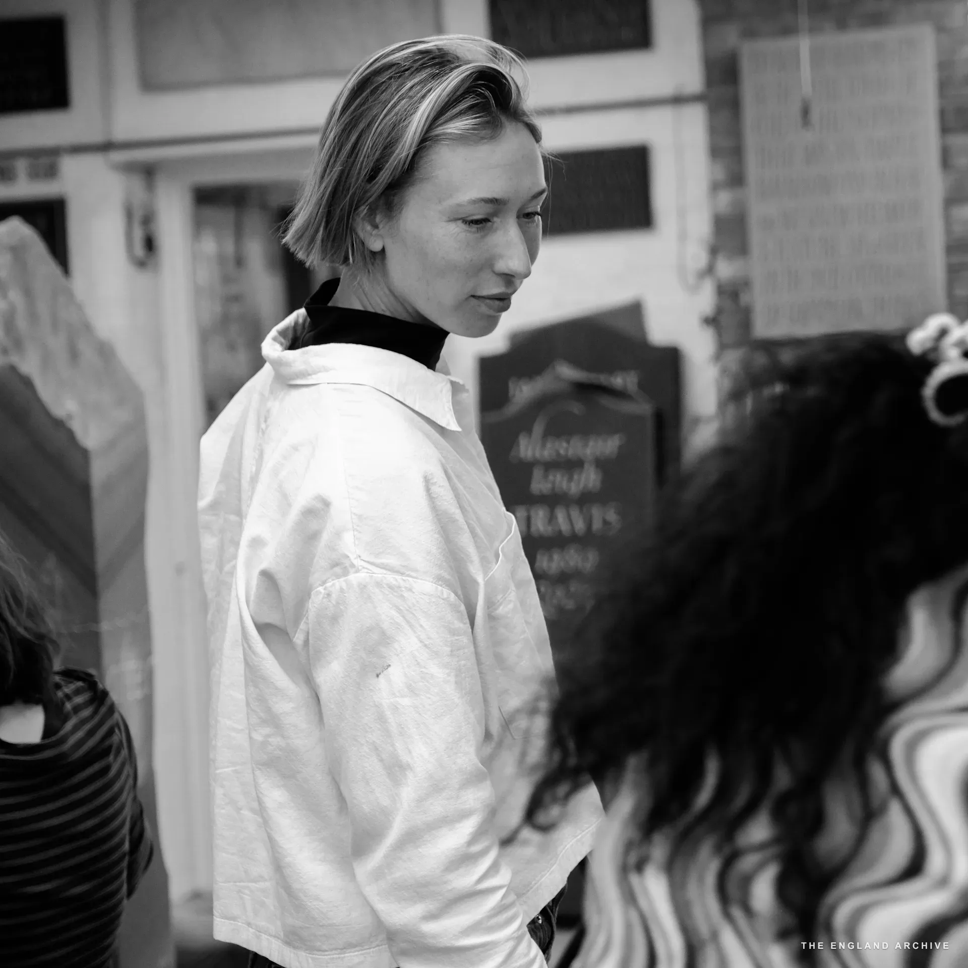 Roxanne Kindersley in profile at the workshop, in a white shirt, listening; another team member with curly hair stands turned away in the foreground; carved slate signs visible on the wall behind.