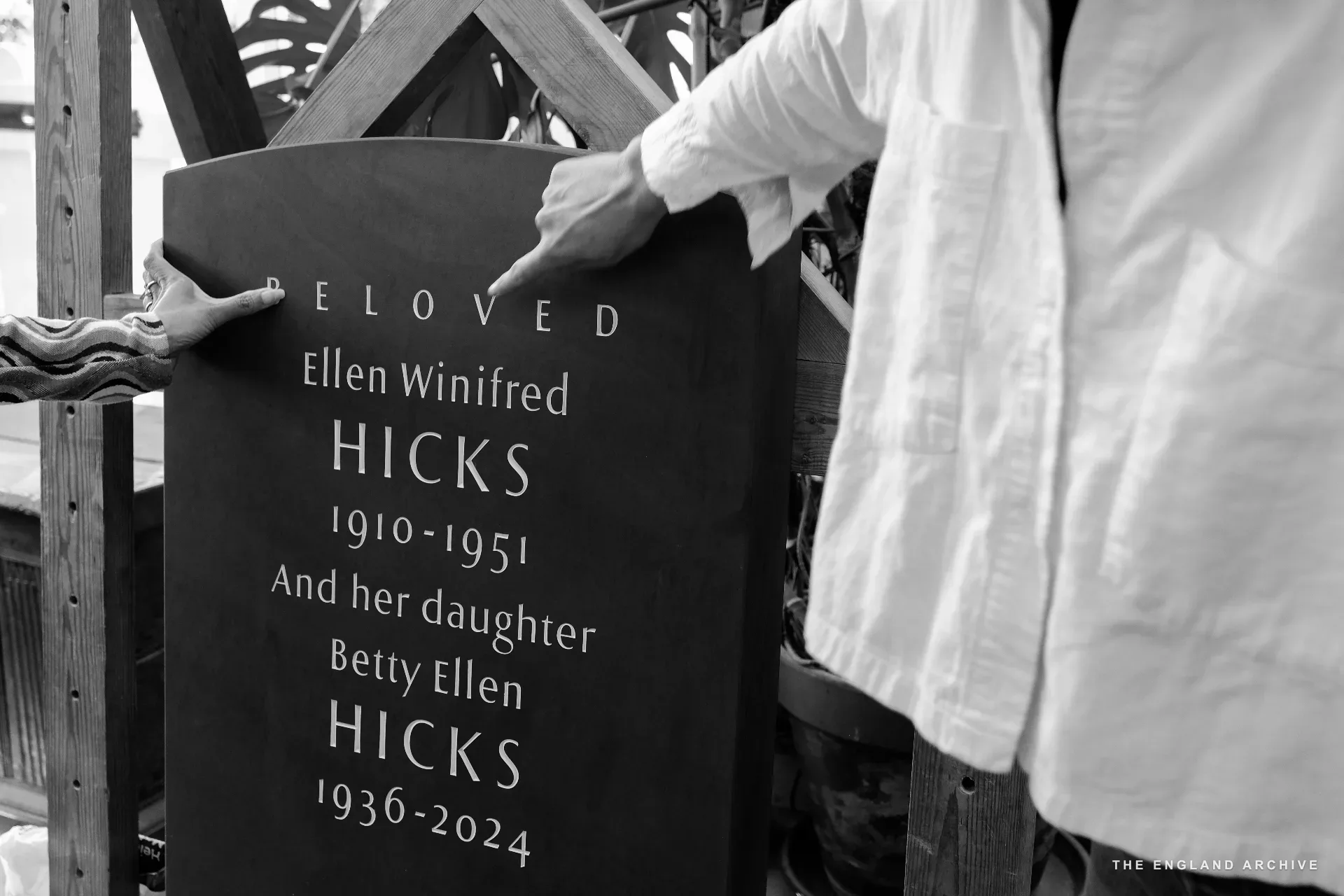 A finished slate memorial stone, dark and freshly cut, reading 'BELOVED Ellen Winifred HICKS 1910-1951 And her daughter Betty Ellen HICKS 1936-2024'. Two hands - one on either side - rest on the stone's edge while it is steadied on its wooden support.