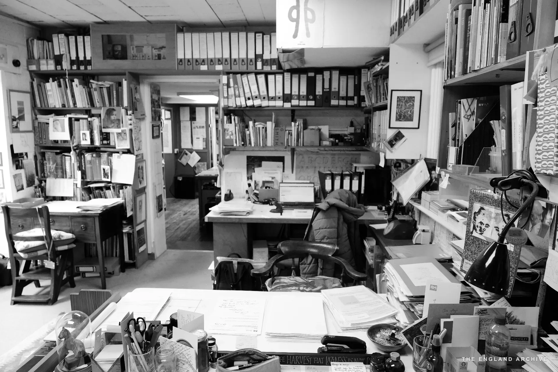 The workshop's back office seen through the doorway: tall shelves of black-spined files filling the walls, a wooden chair drawn up to a paper-strewn desk, books and finished work stacked on every surface.