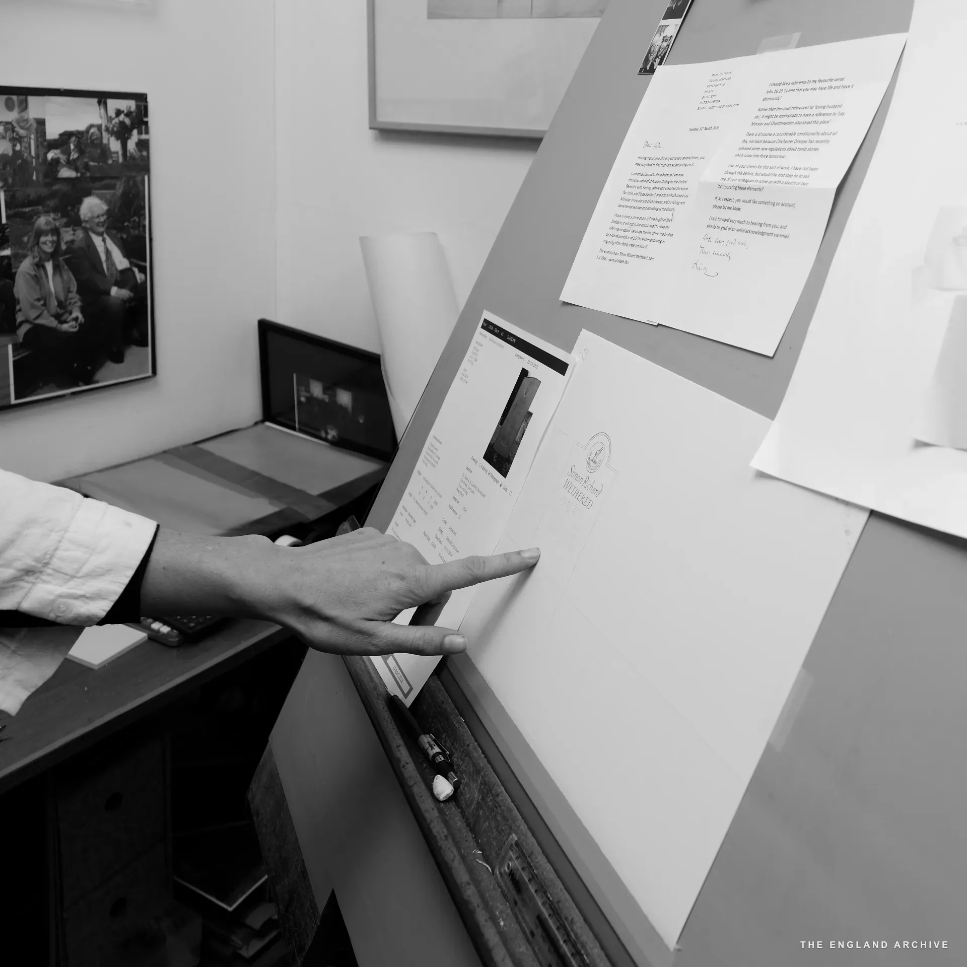 A hand pointing at the working sketch of a memorial stone for Simon Richard Wethered, pinned to the workshop's drafting board; a long client letter pinned alongside, a framed photograph of two figures in a garden visible to the left.