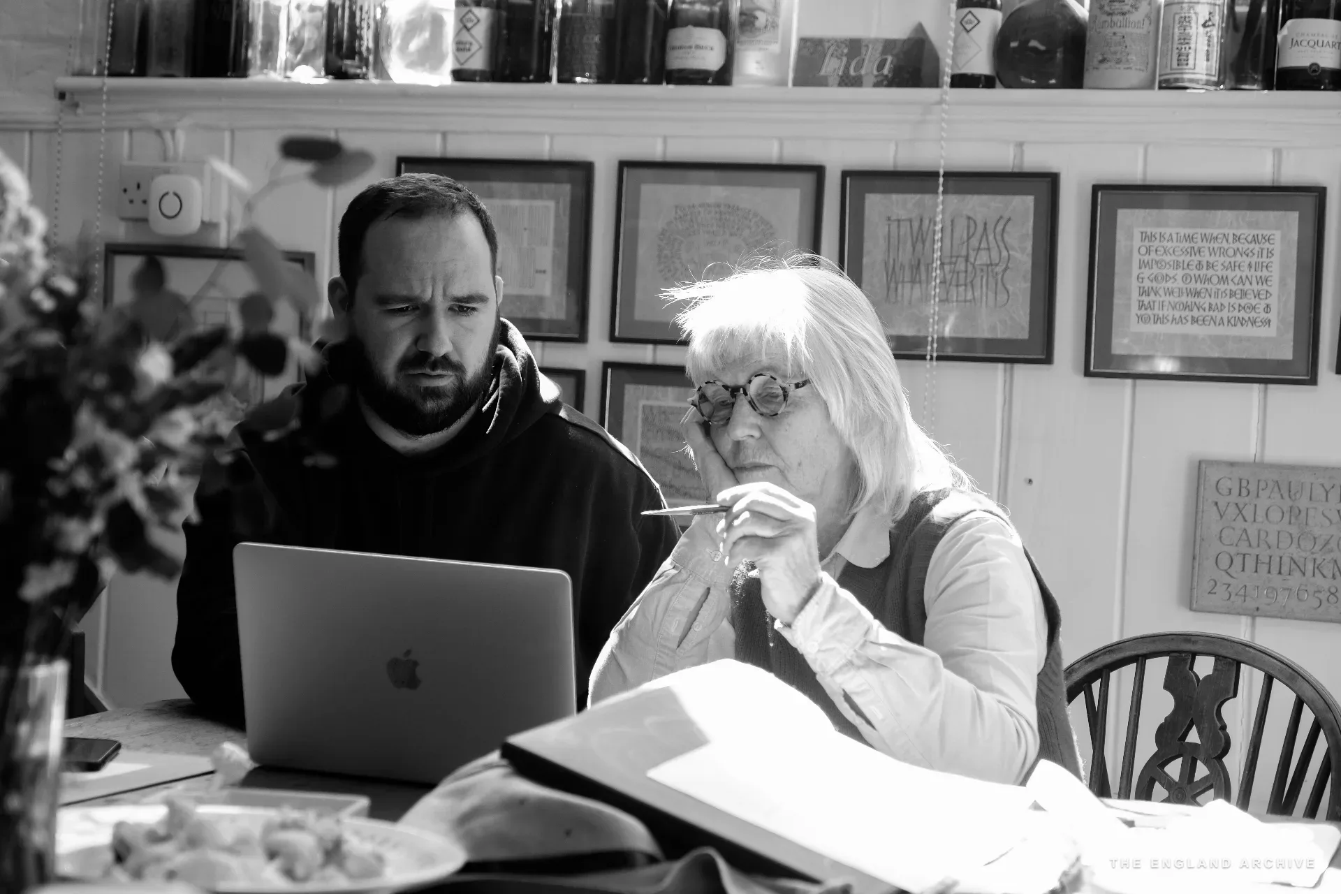 Lida Kindersley and a younger bearded man with glasses sit side by side at the front-room table, the man working at a laptop, Lida holding her glasses in one hand mid-thought, looking down at an open book between them. Bottles of wine on the high shelf above; framed letter samples crowd the wall.