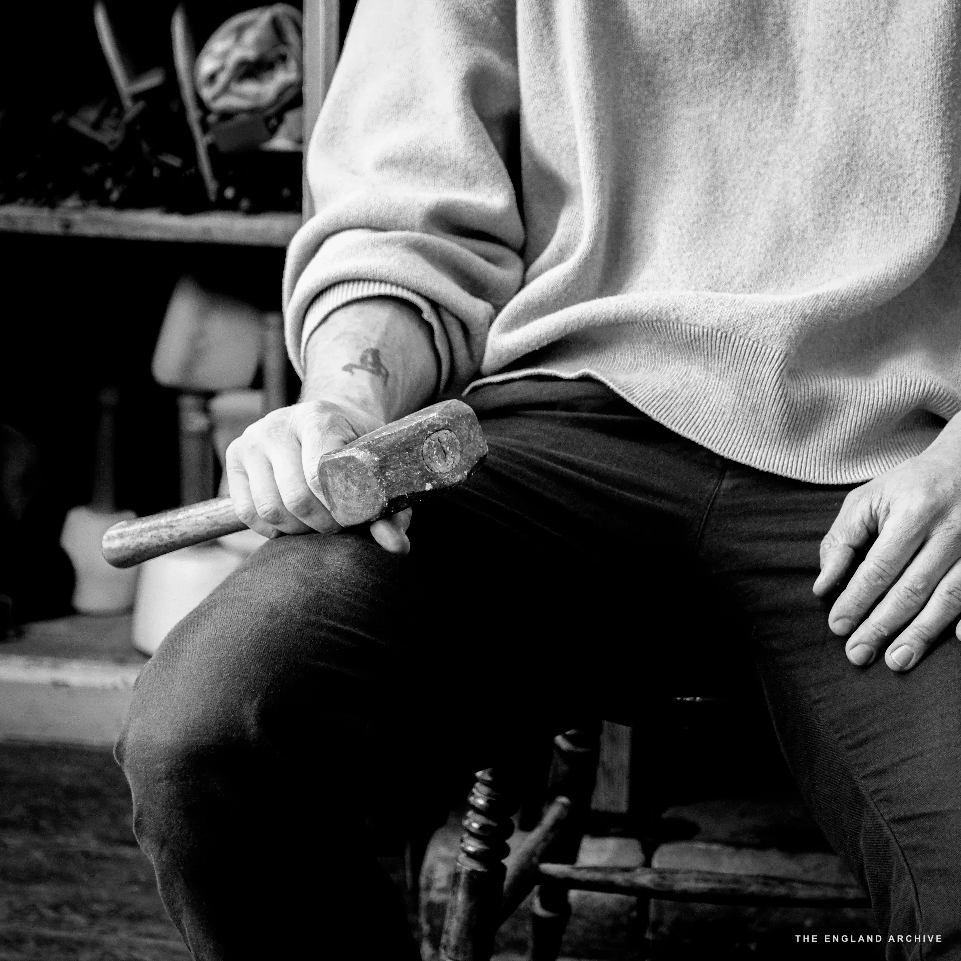 Close on a man's lap and torso: pale jumper, dark jeans, a stone-cutting hammer held loosely in the left hand. A small tattoo on the inner forearm. The workshop's chisel-rack pigeonholes are softly out of focus behind.