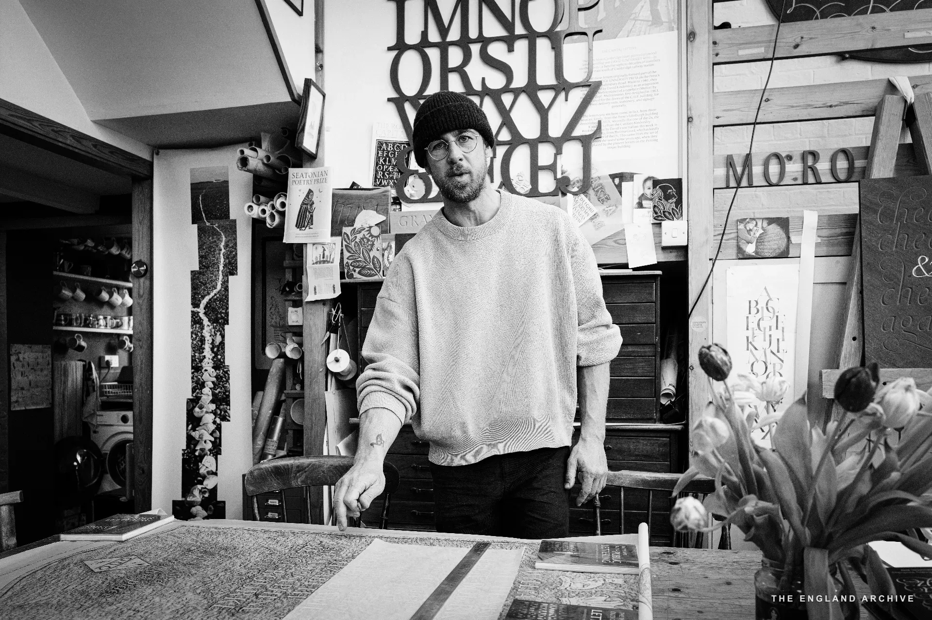 A man in a beanie and cream sweatshirt standing in the front room near the table, with workshop letter-cuts and pinned reference photos on the wall behind, a vase of tulips on the table and the workshop's painted MORO sign visible to the right.